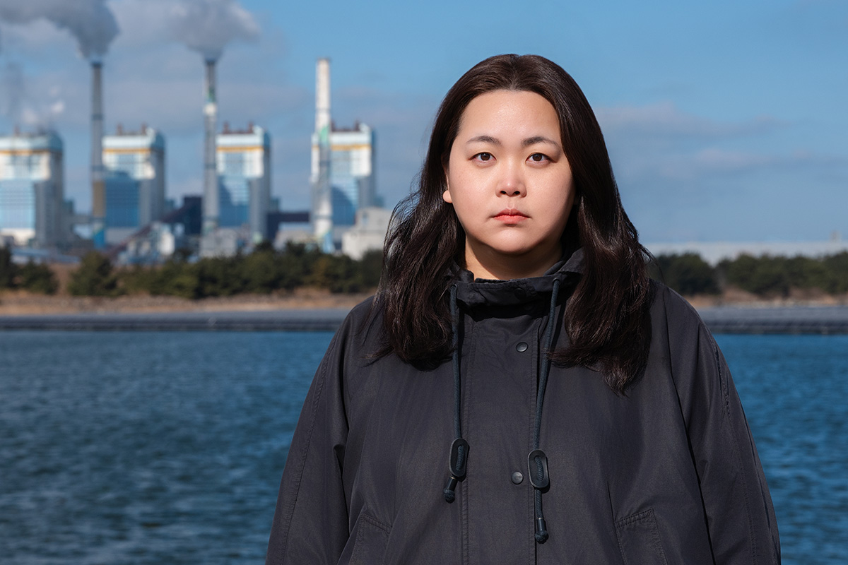 Personne debout devant une usine industrielle avec cheminées fumantes en arrière-plan, près de l'eau.