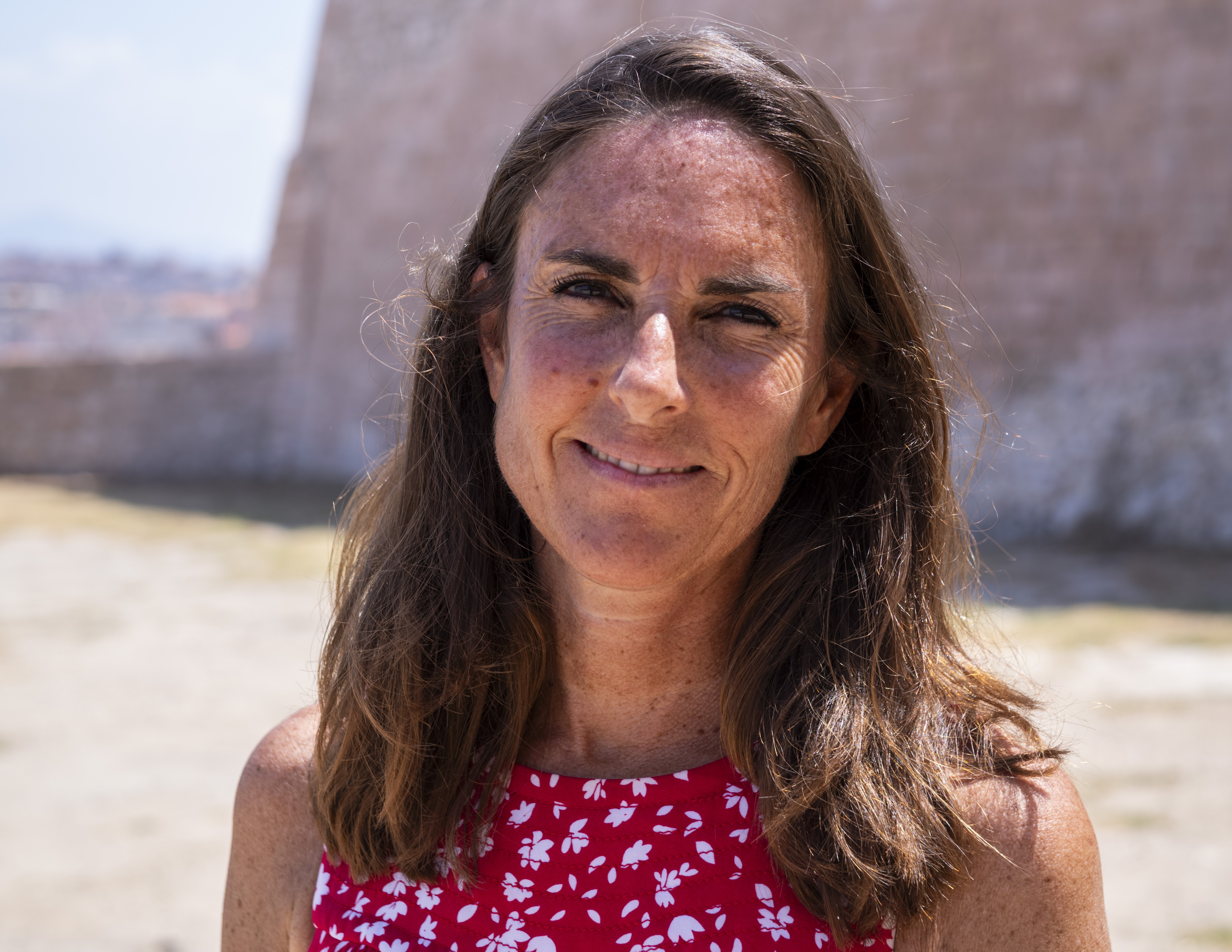 Femme souriante devant un mur en pierre, vêtue d'un haut rouge, cheveux longs.