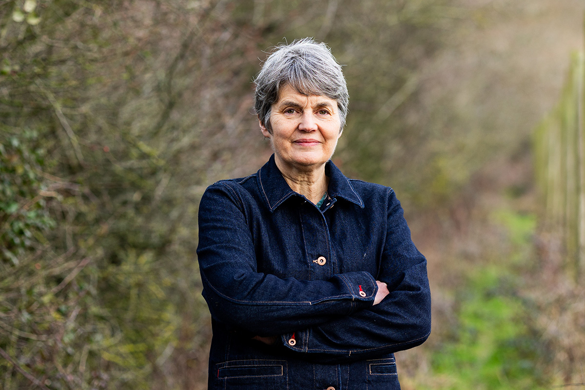 Femme aux cheveux gris dans un jardin, bras croisés, veste sombre, fond végétal flou.