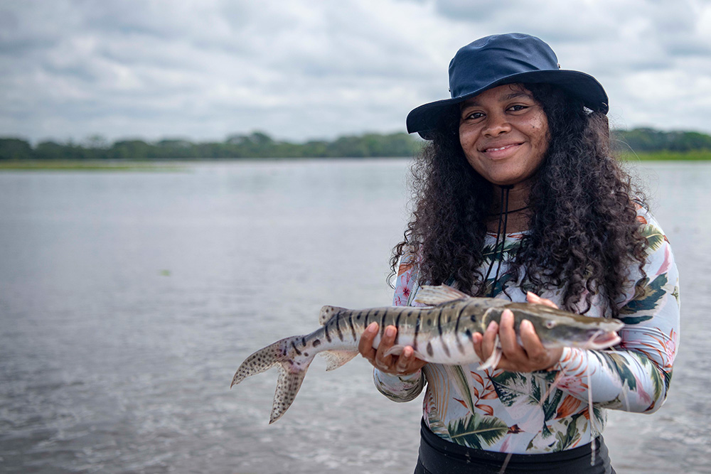 Femme souriante avec chapeau tenant un poisson devant un lac sous un ciel nuageux.