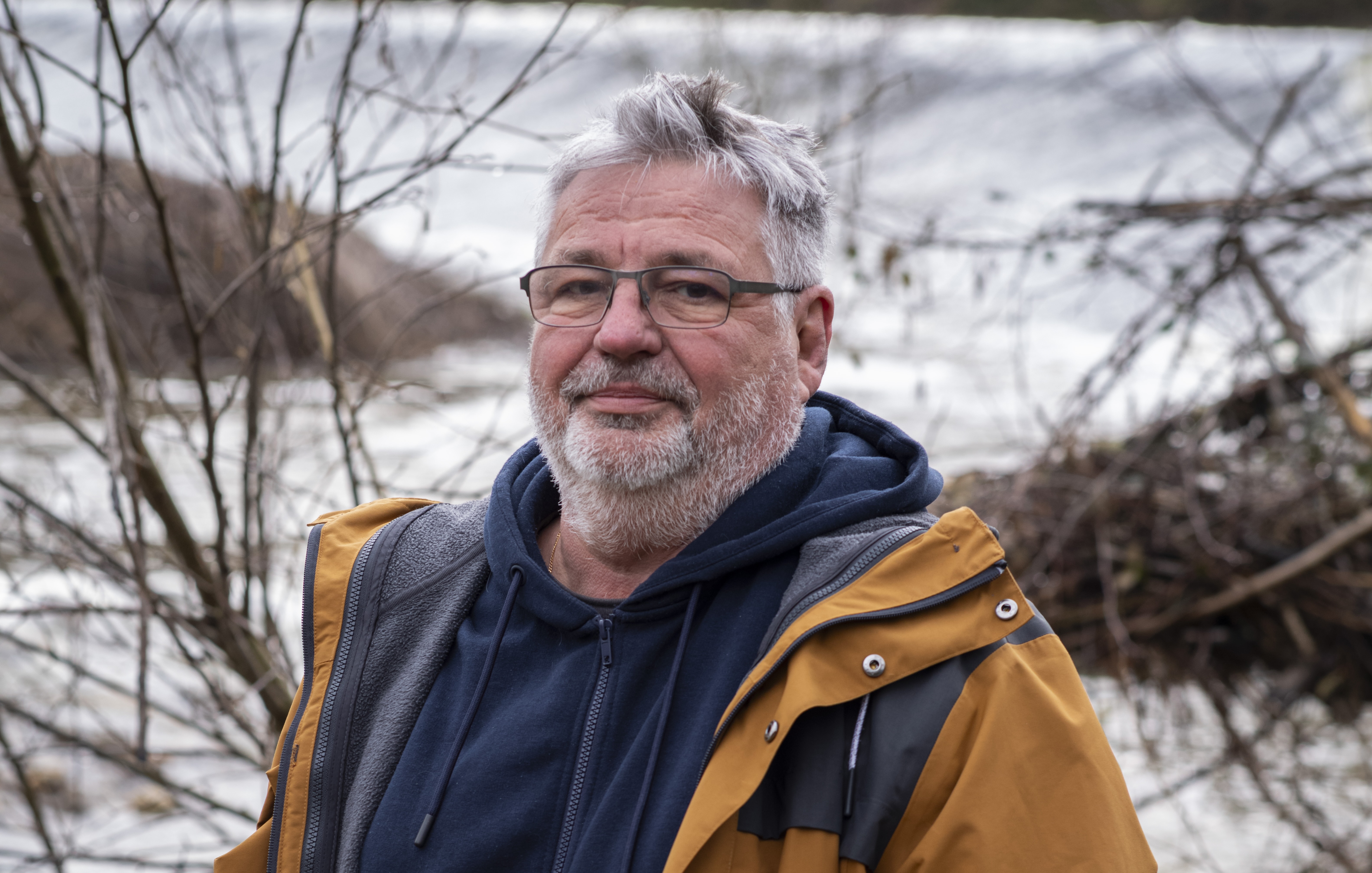 Homme avec barbe grise en extérieur, portant une veste jaune devant un paysage fluvial.