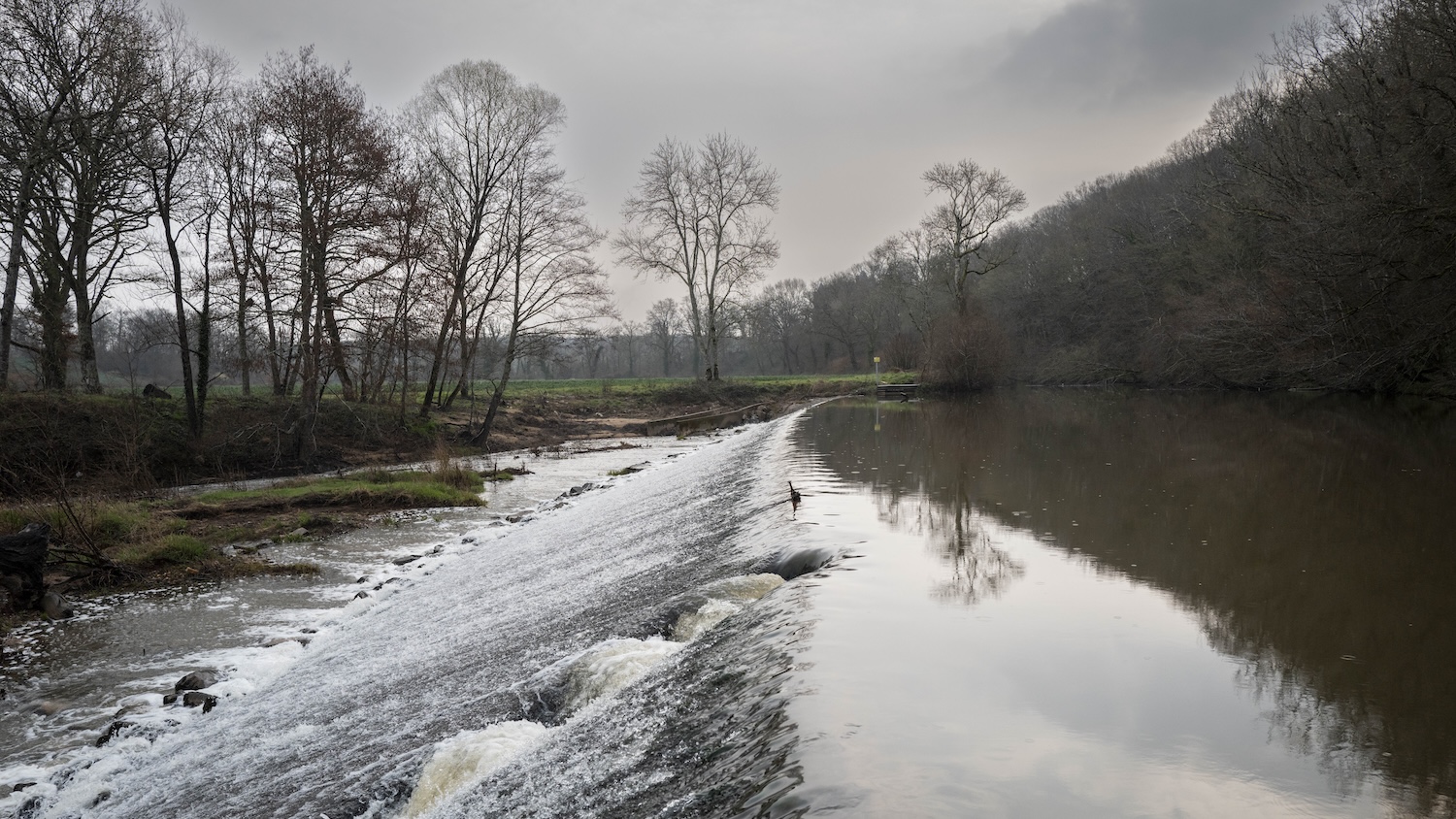Rivière avec cascade entourée d'arbres en hiver sous un ciel nuageux.