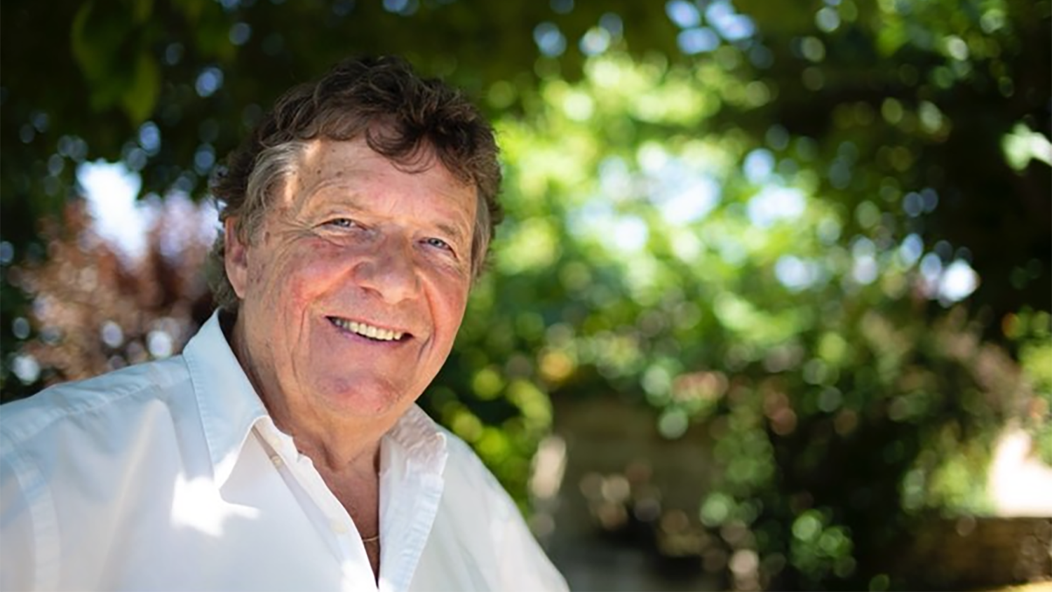 Homme souriant en chemise blanche devant des arbres, éclairé par le soleil.