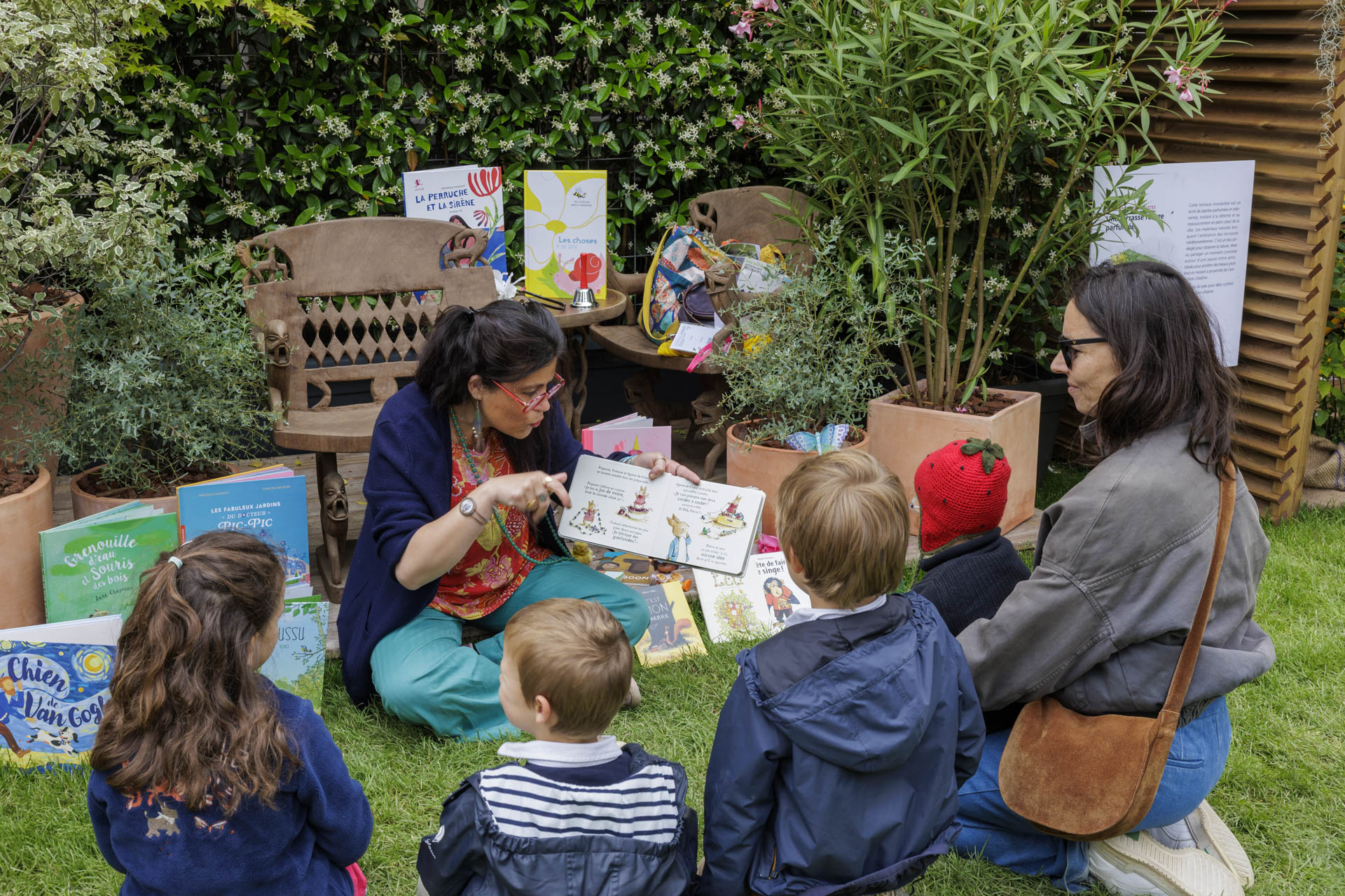 Femme lisant des histoires à des enfants assis dans un jardin entouré de livres.