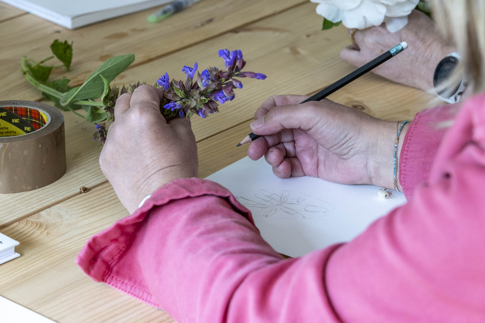 Personne en chemise rose dessinant une fleur violette sur une feuille de papier.