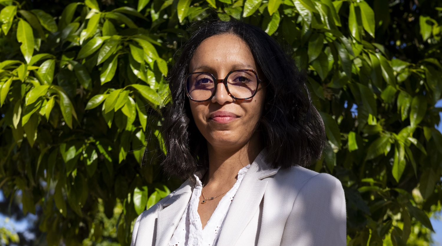 Femme avec lunettes, cheveux noirs, portant un blazer, debout devant un arbre feuillu.