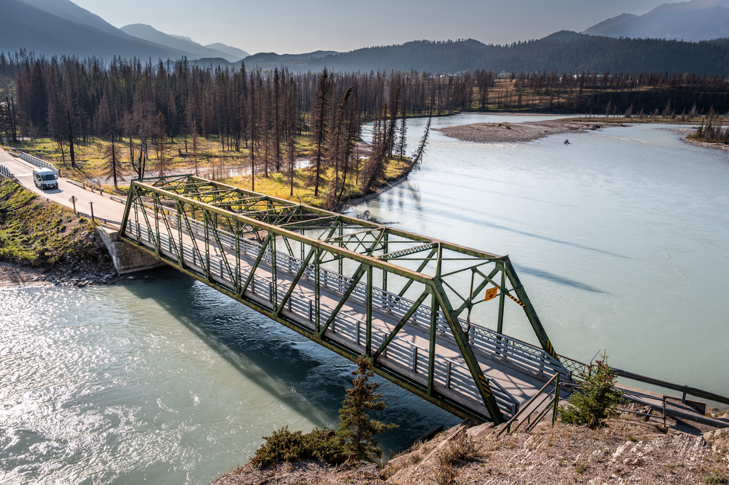 Pont métallique traversant une rivière avec voiture et paysage forestier en arrière-plan.