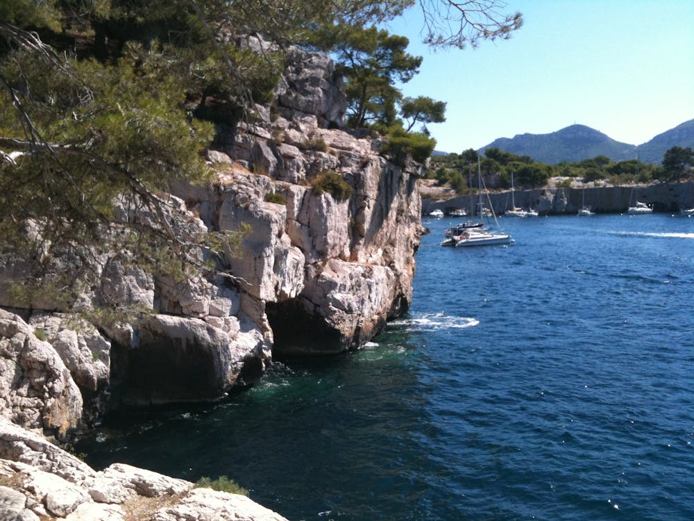 Falaises rocheuses avec pins surplombant une mer bleue, bateaux au loin, journée ensoleillée.