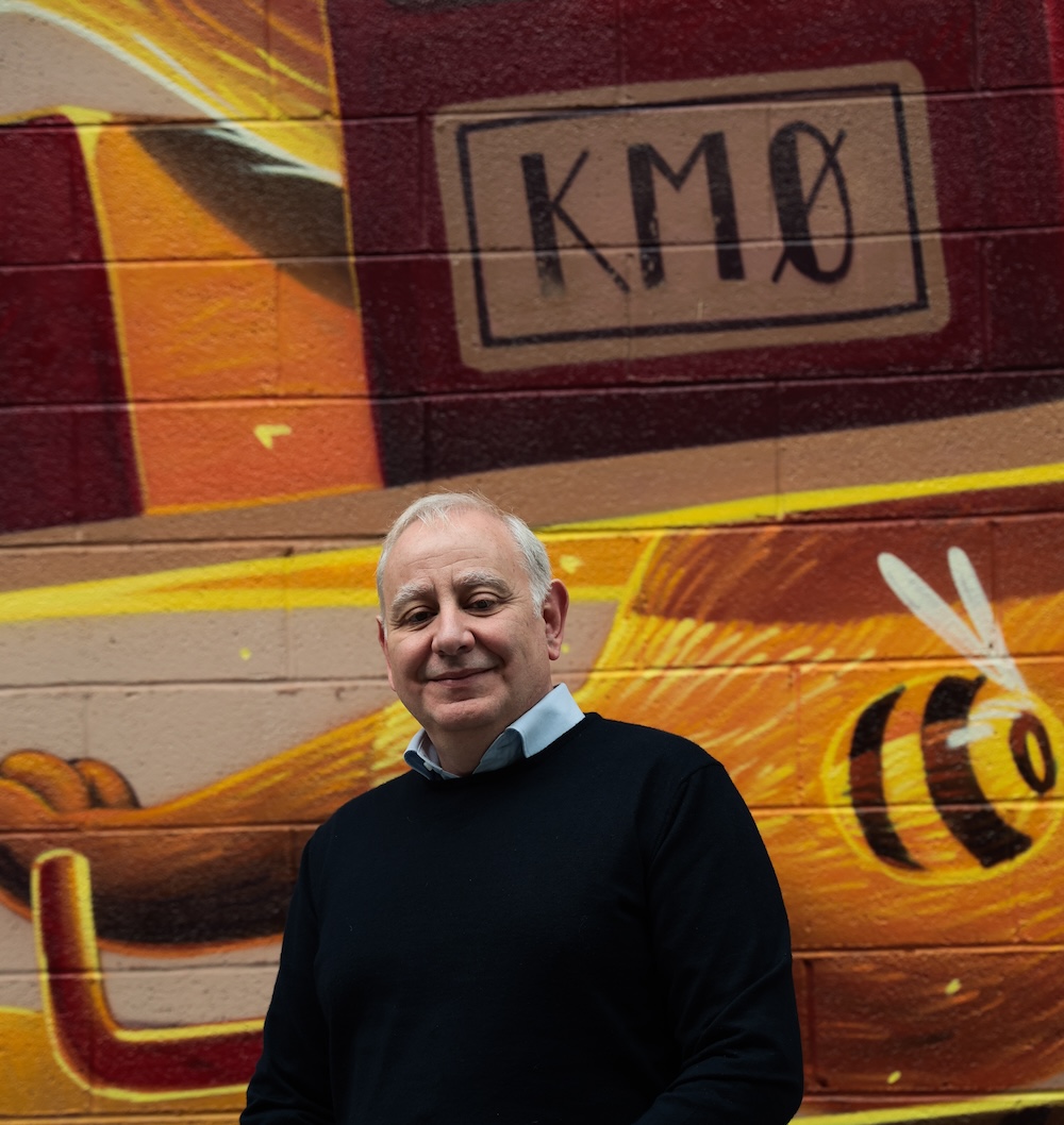 Homme souriant en pull noir devant une fresque murale colorée avec une abeille.