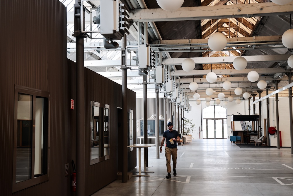 Un homme marche dans un grand espace industriel avec des structures métalliques et des fenêtres.