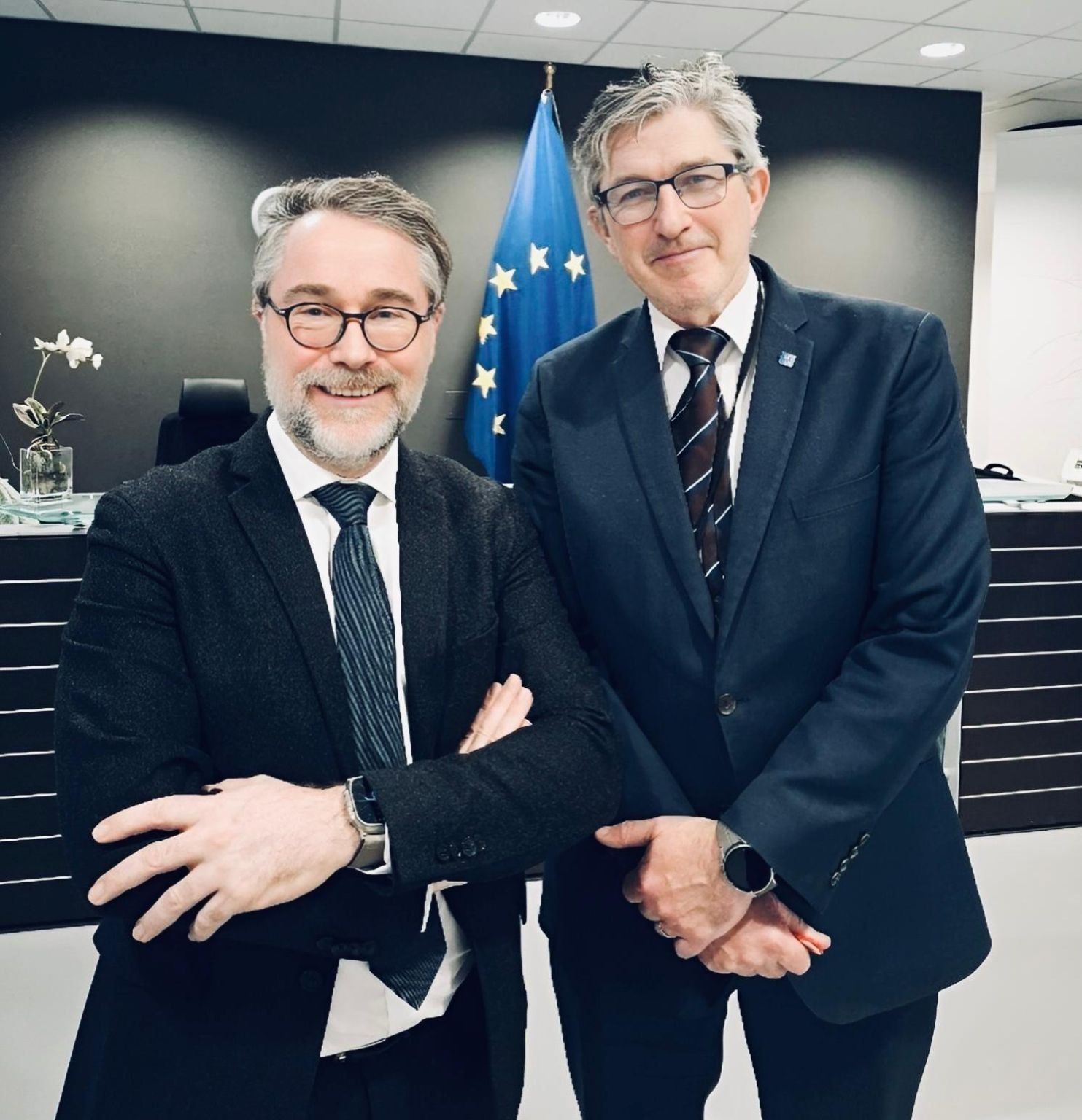 Deux hommes en costume souriants devant un drapeau de l'Union européenne.