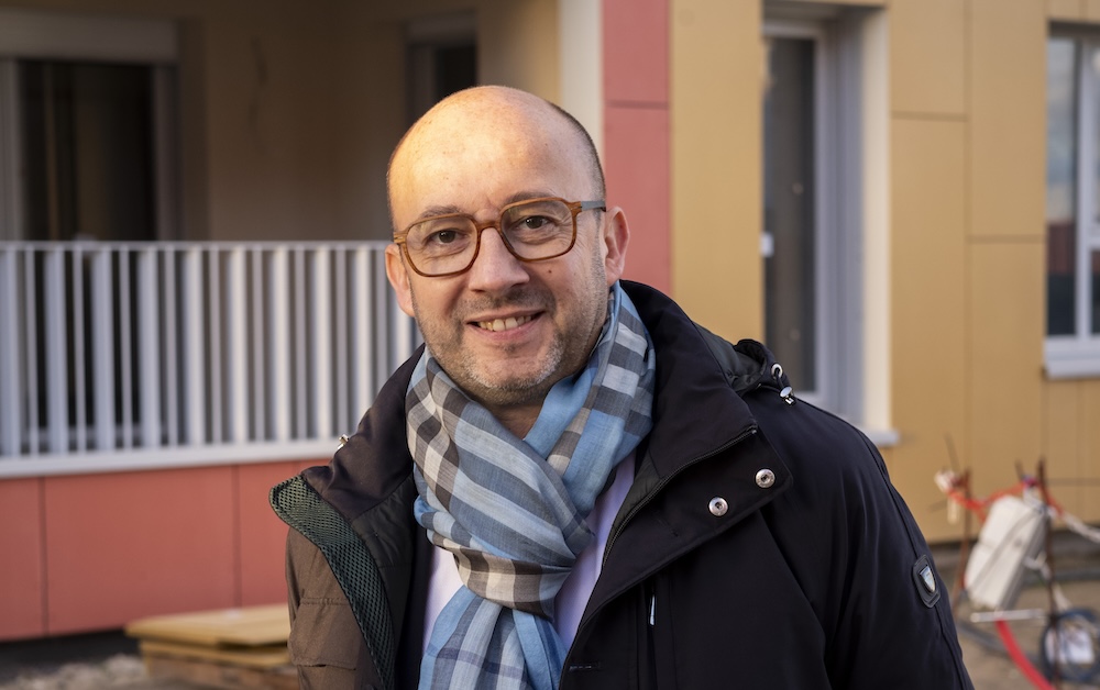 Homme souriant portant une écharpe et des lunettes devant un bâtiment coloré.