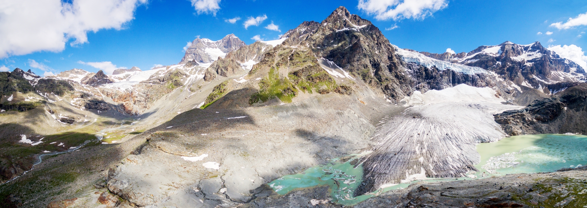 Paysage alpin avec montagnes enneigées, vallée rocheuse et lac turquoise sous un ciel bleu.