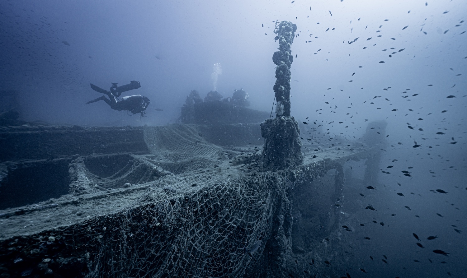 Plongeur explorant une épave sous-marine entourée de poissons et de filets.