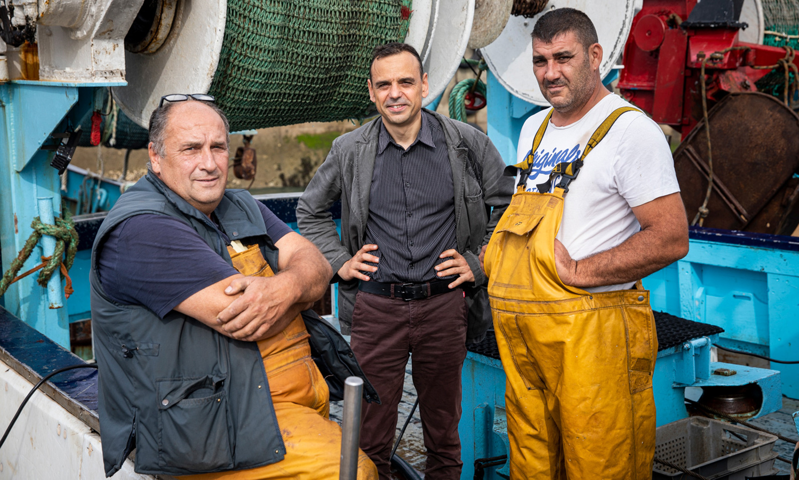 Trois hommes posent sur un bateau de pêche avec équipements et filets.