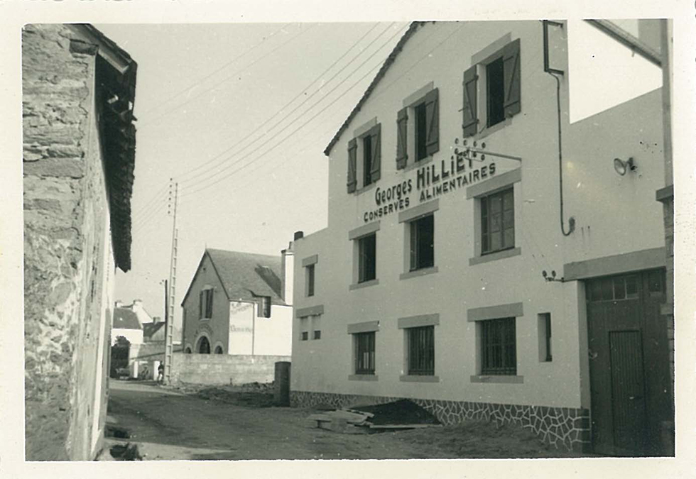 Bâtiment industriel ancien avec écriteau "Moulin" dans une rue étroite, maisons avoisinantes.