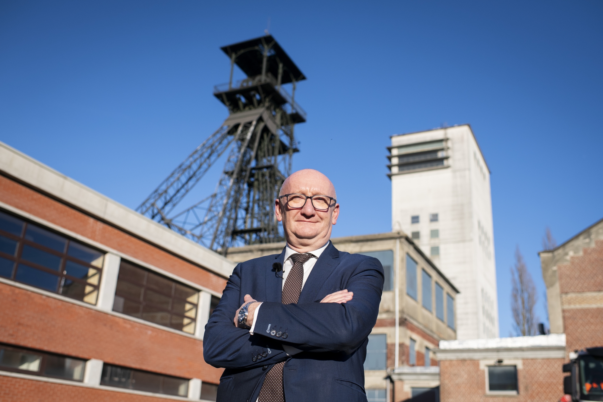 Homme en costume debout devant un ancien site industriel avec structure métallique et bâtiments en briques.