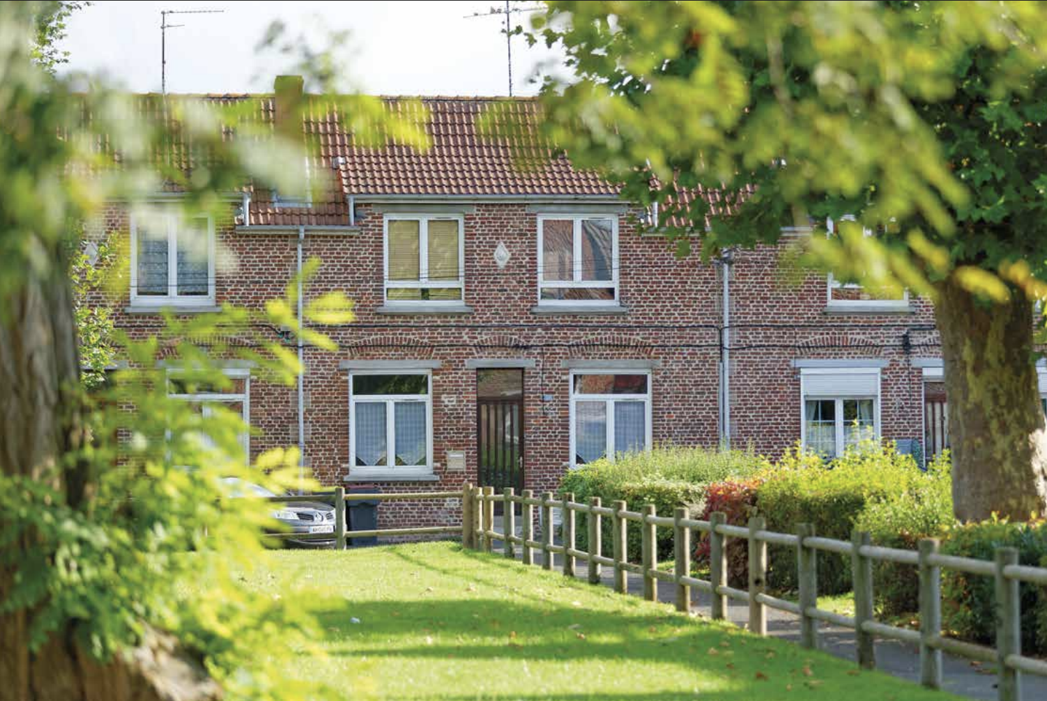 Maison en briques rouges avec jardin vert et allée bordée d'arbres et de buissons.