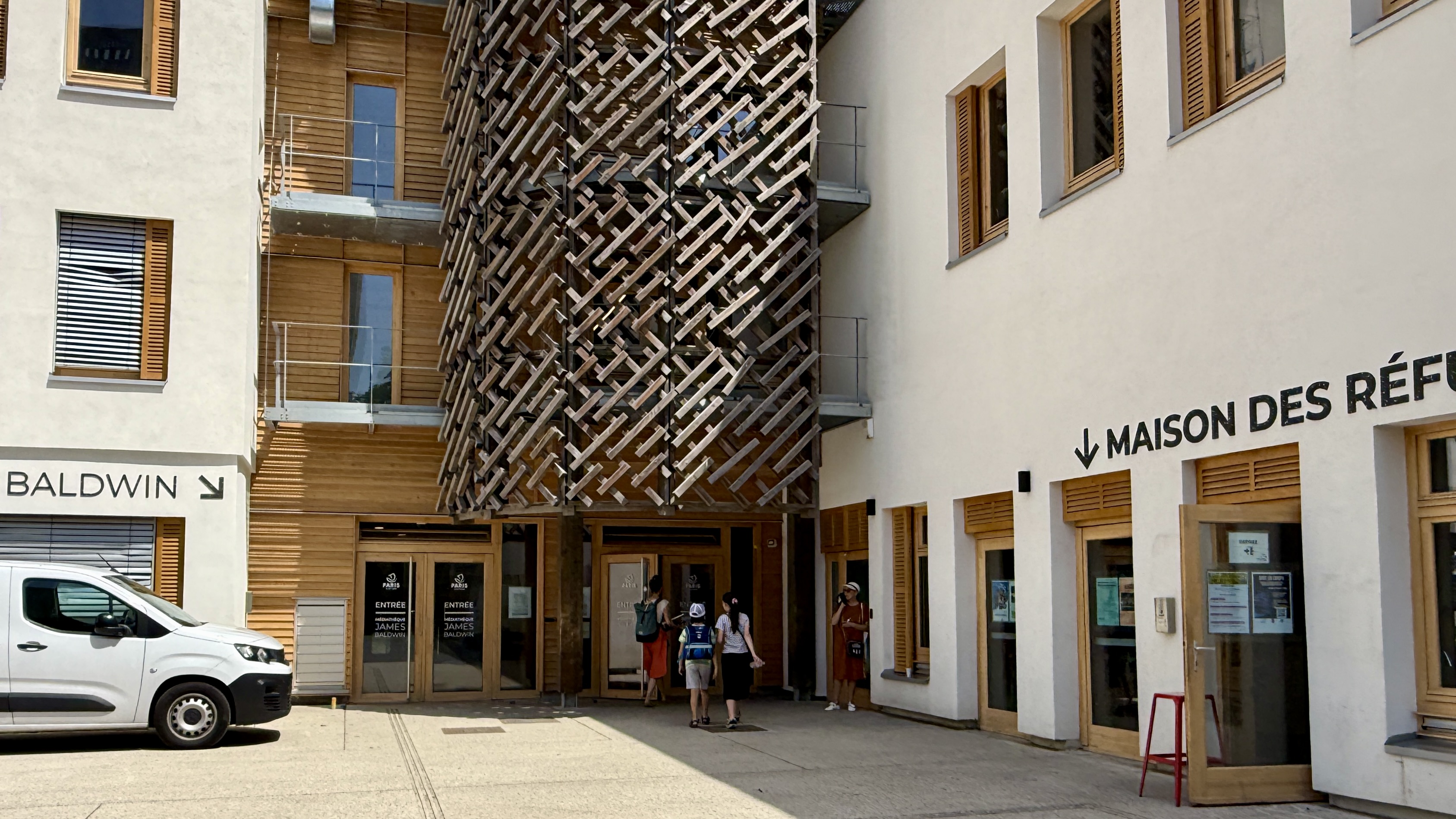 Façade de la Maison des Réfugiés avec des personnes entrant et une voiture garée devant.