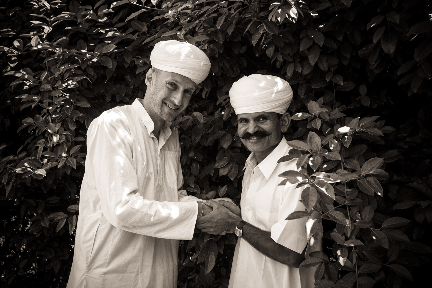 Deux hommes souriants en tenues traditionnelles blanches se serrent la main devant des buissons.