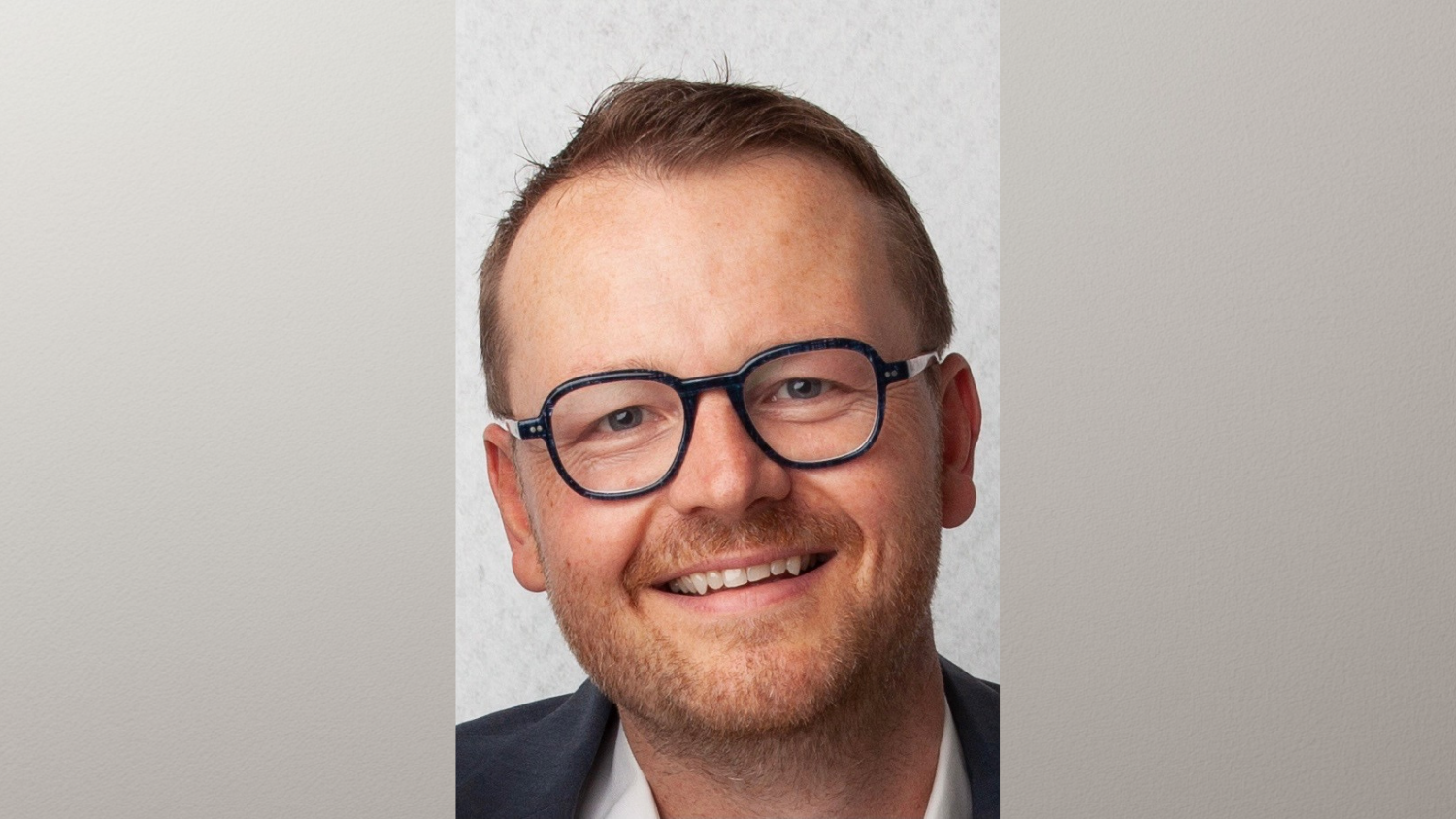 Homme souriant portant des lunettes et une veste, fond clair et neutre.