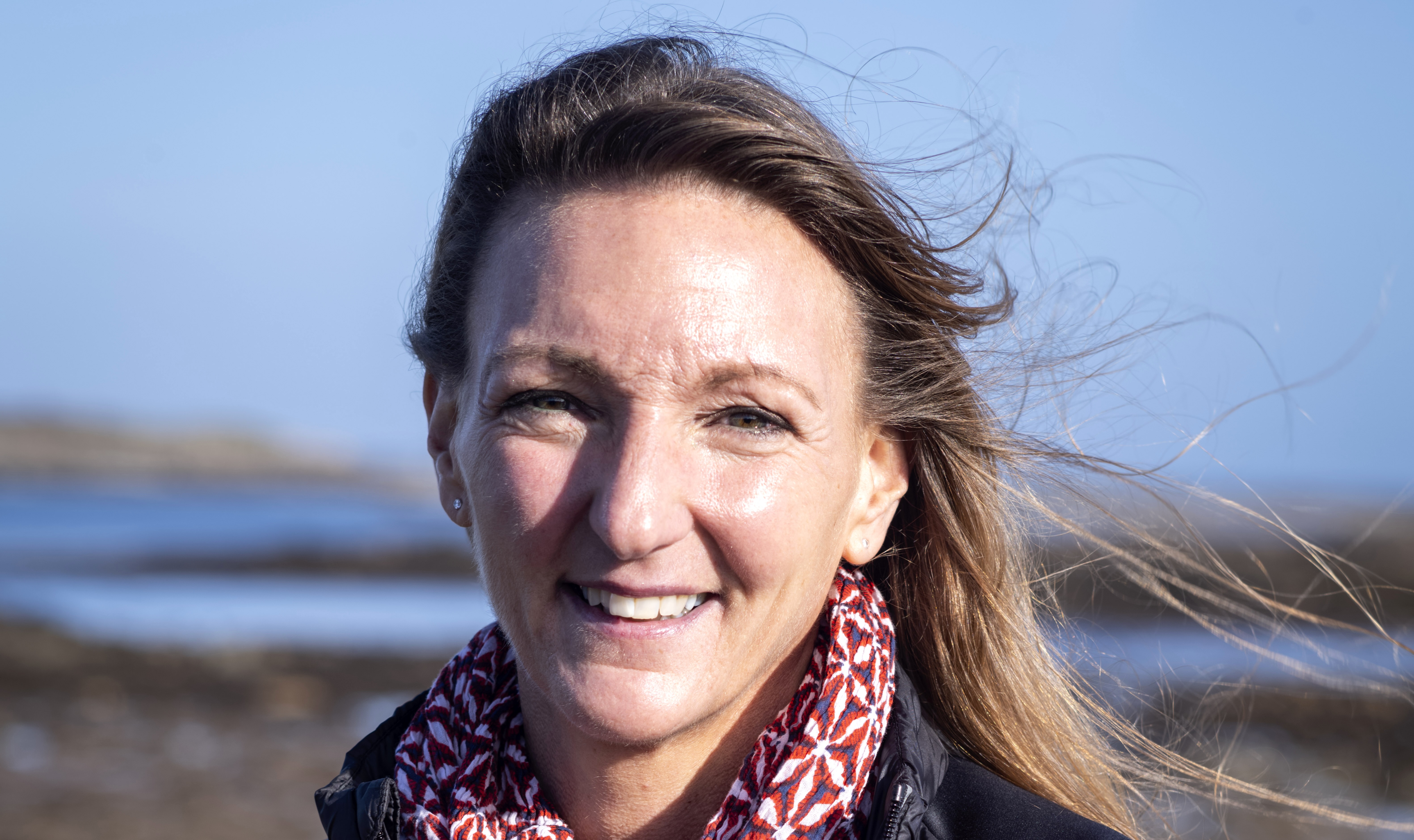 Femme souriante en extérieur avec un foulard rouge, ciel bleu et paysage marin en arrière-plan.