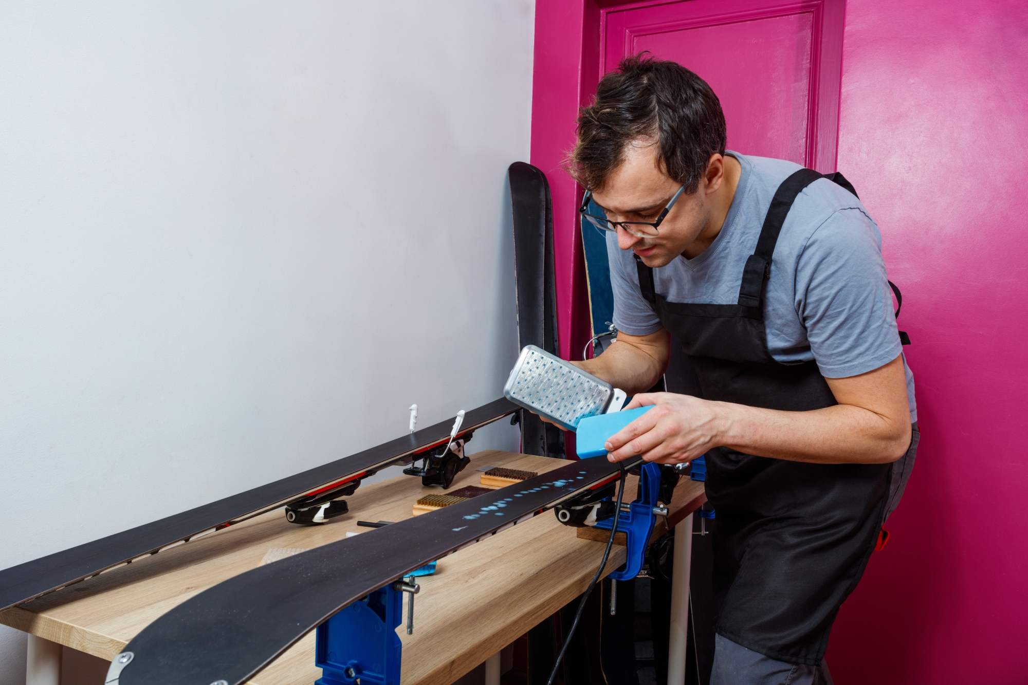 Un homme entretient des skis dans un atelier avec un mur rose en arrière-plan.