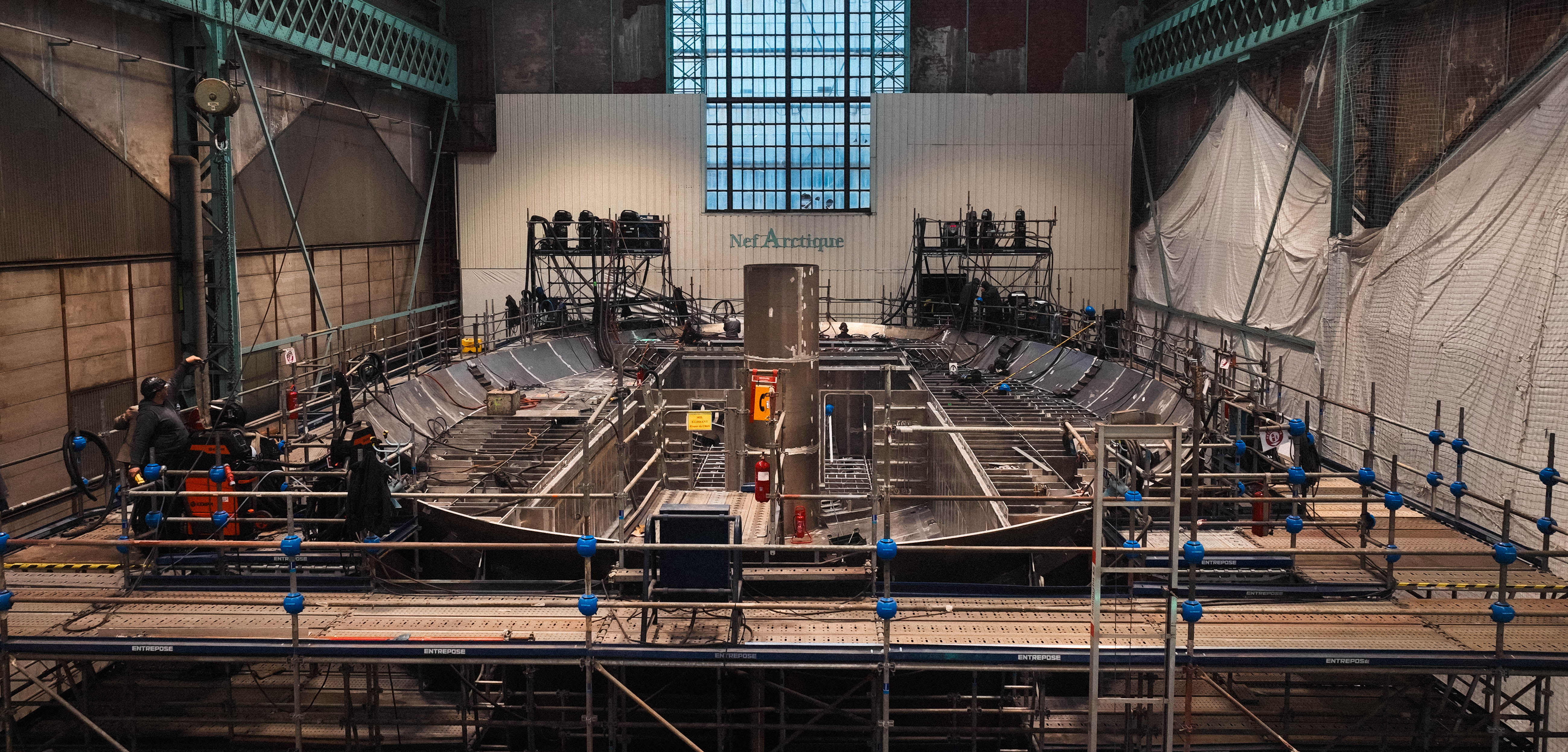 Travaux de construction à l'intérieur d'un grand bâtiment industriel avec échafaudages et structures métalliques.