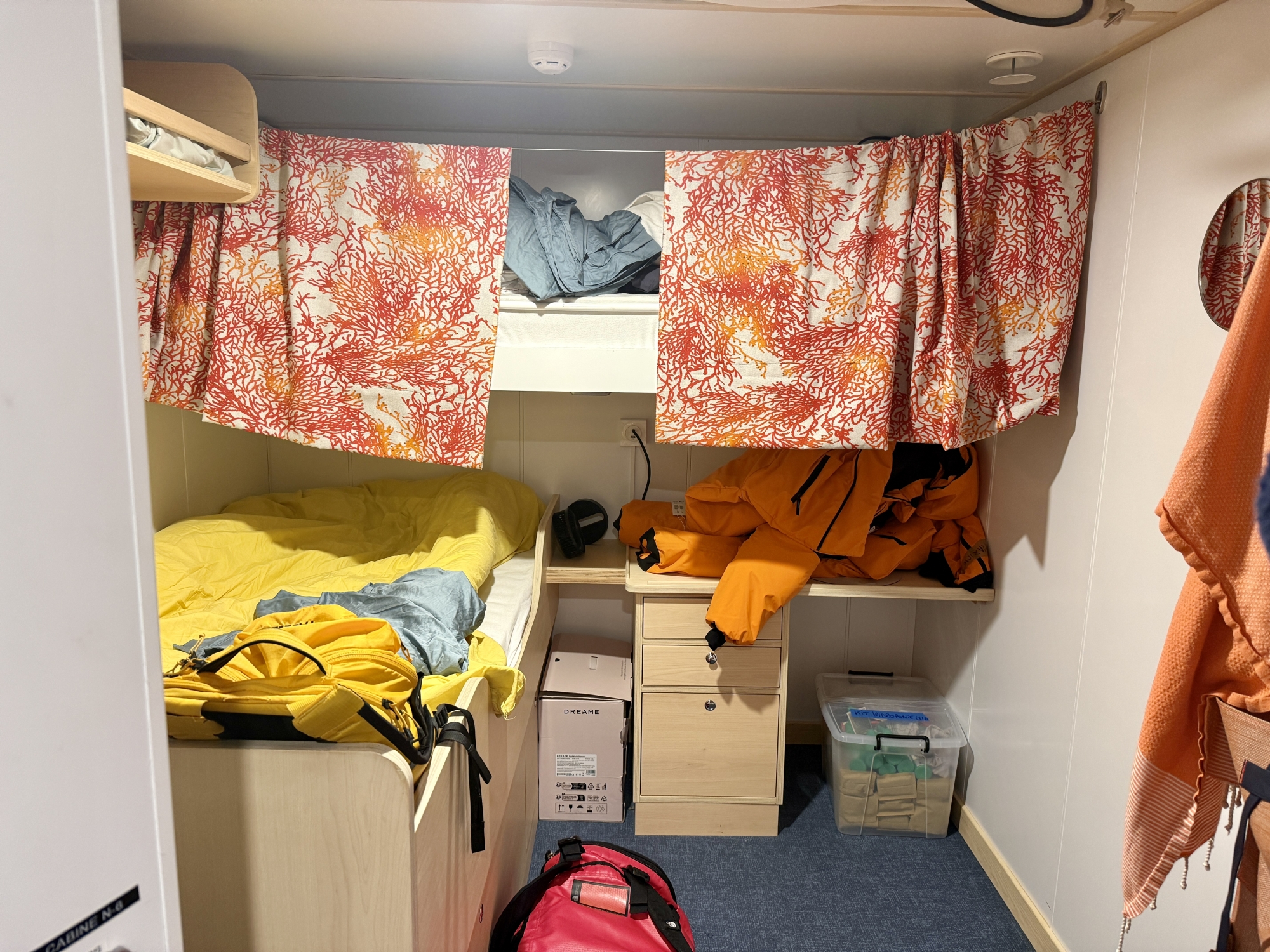 Petit espace avec lit superposé, rideaux colorés, vêtements et boîte de rangement sous le bureau.