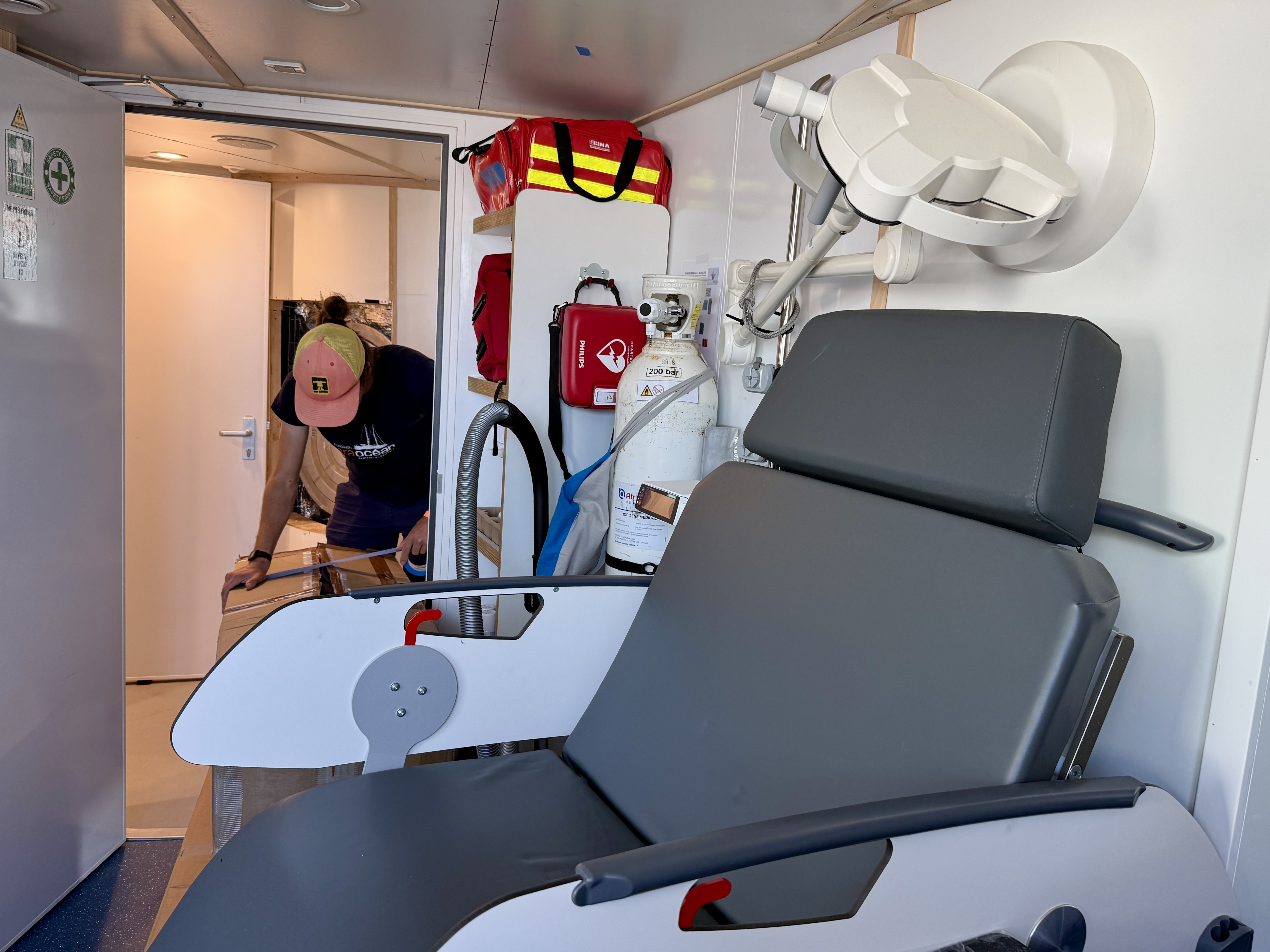 Intérieur d'une infirmerie avec équipement médical et siège, personne entrant par la porte arrière.