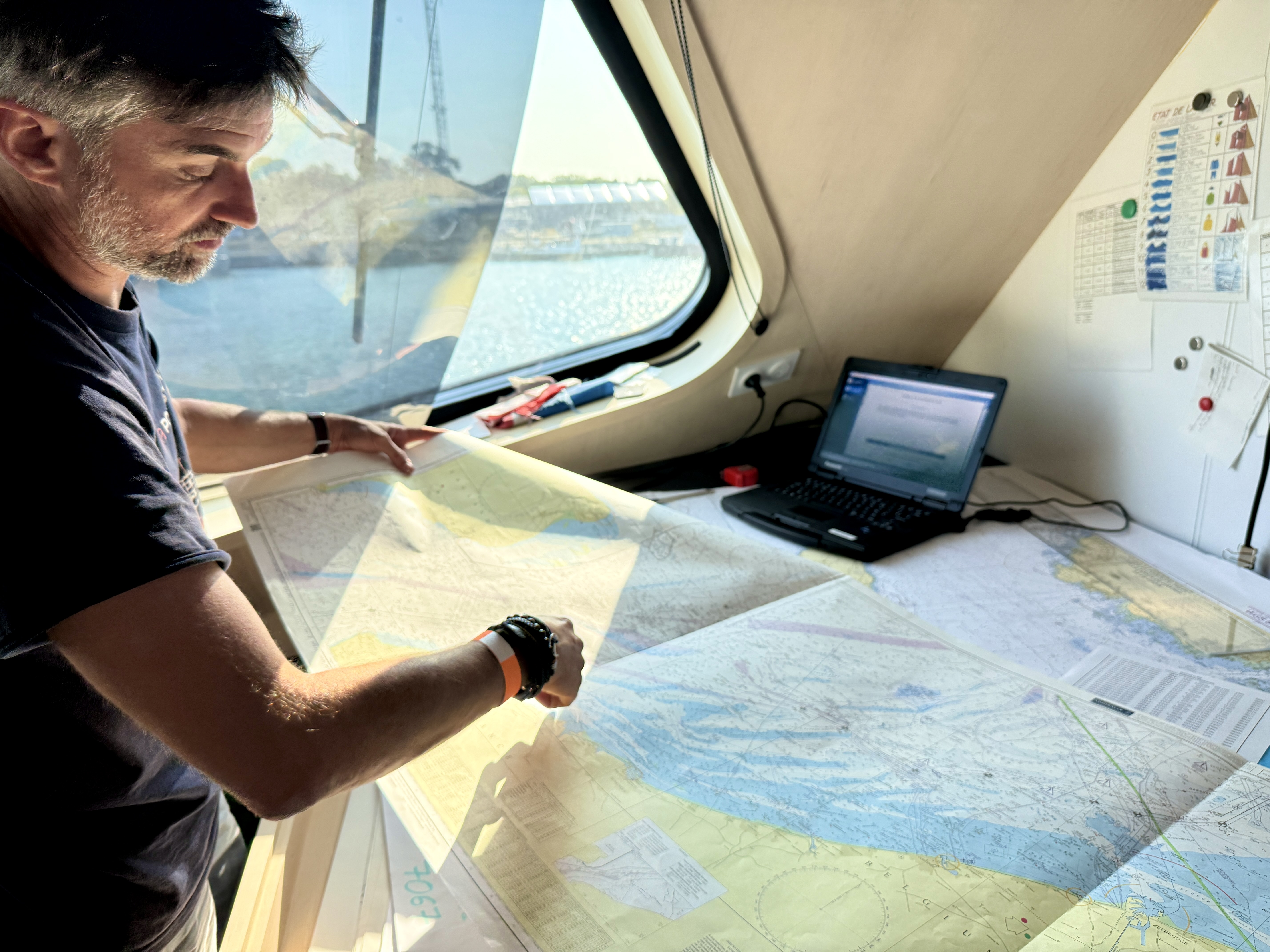 Un homme consulte des cartes maritimes dans une cabine de bateau ensoleillée.