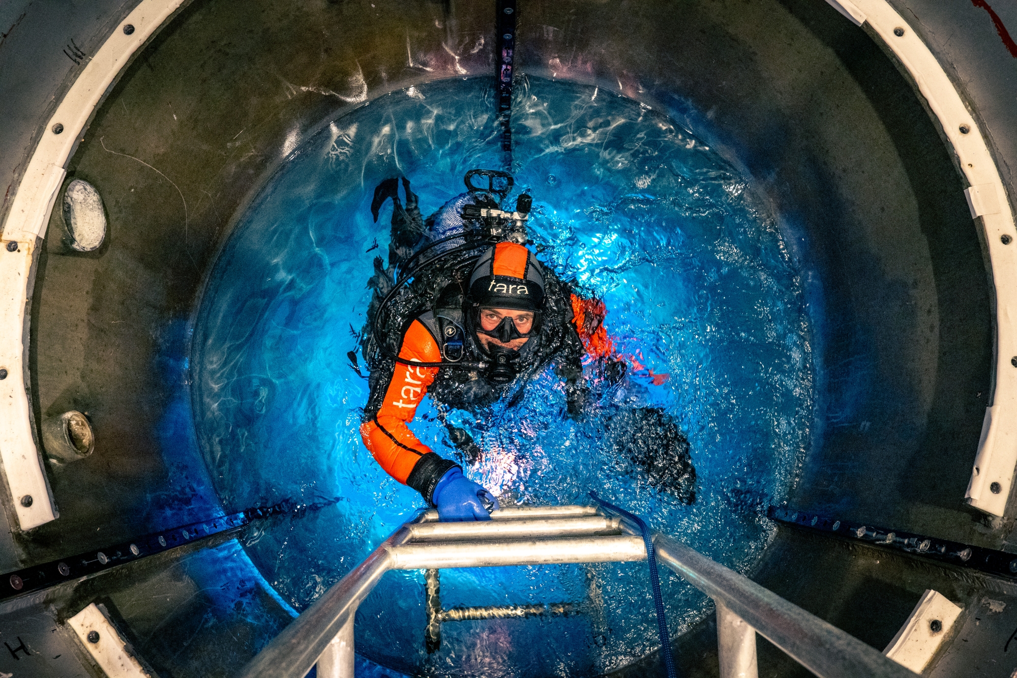 Plongeur en combinaison orange dans un bassin émergeant vers une échelle métallique sous l'eau.