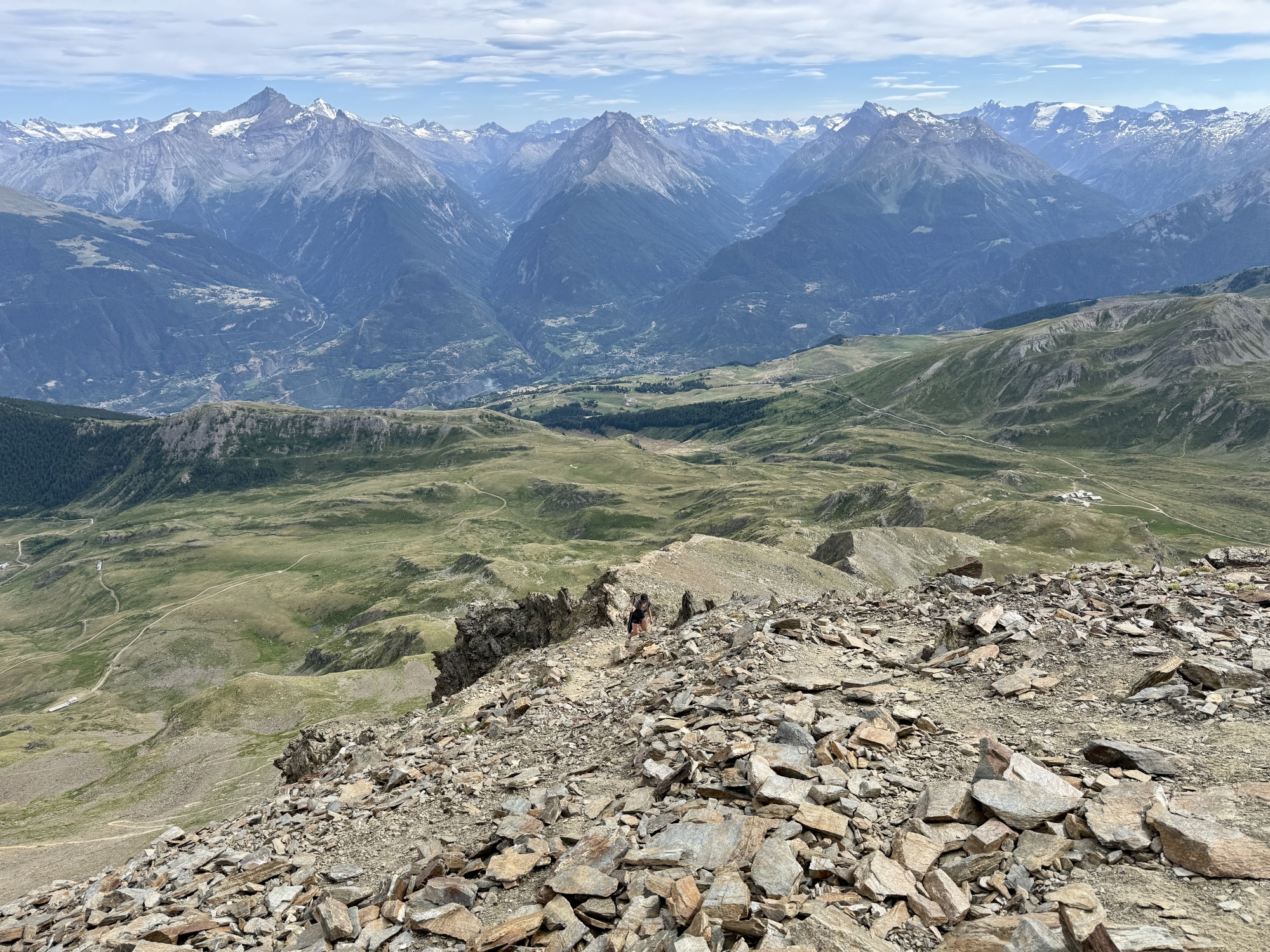 Paysage montagneux avec sentier rocheux, vue sur vallée et montagnes enneigées à l'horizon.