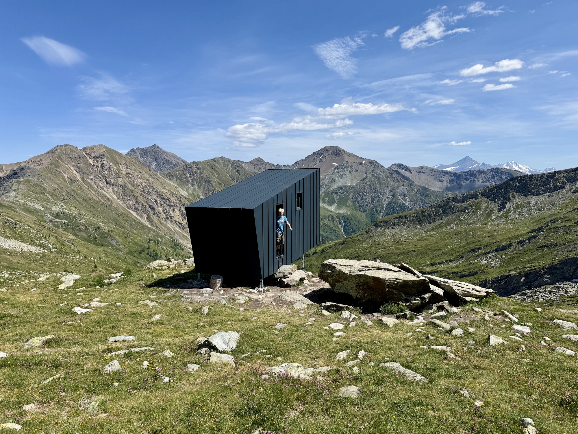 Cabane moderne en montagne avec une personne dans l'entrée, vue sur chaîne de montagnes.