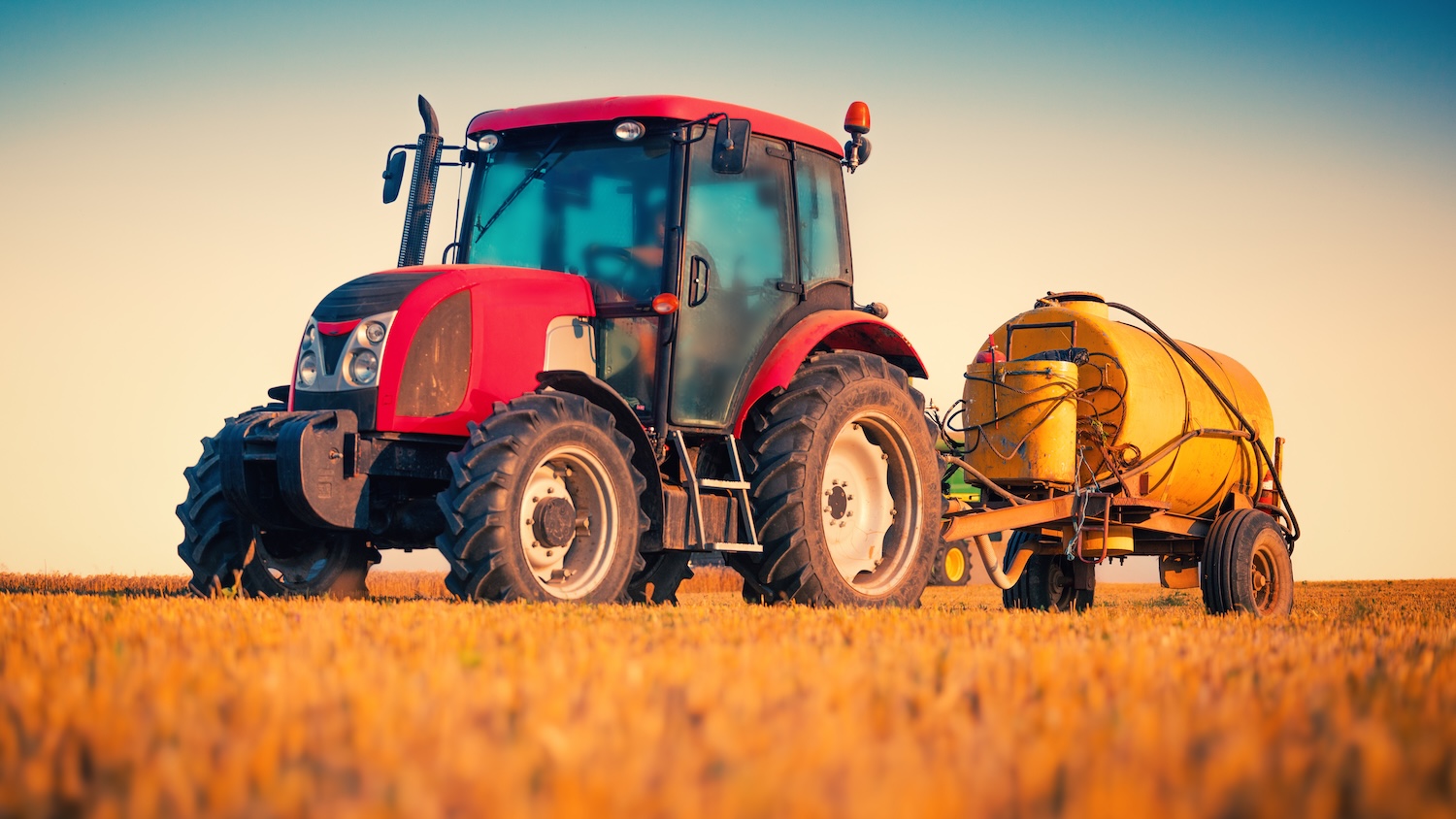 Tracteur rouge dans un champ, tirant une citerne jaune sous un ciel bleu.
