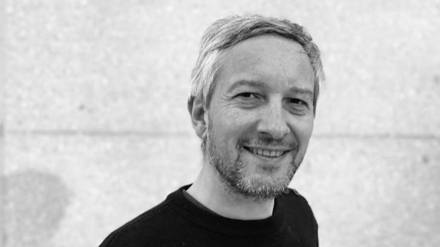 Un homme souriant avec une barbe, vêtu d'un pull noir, sur fond gris clair.