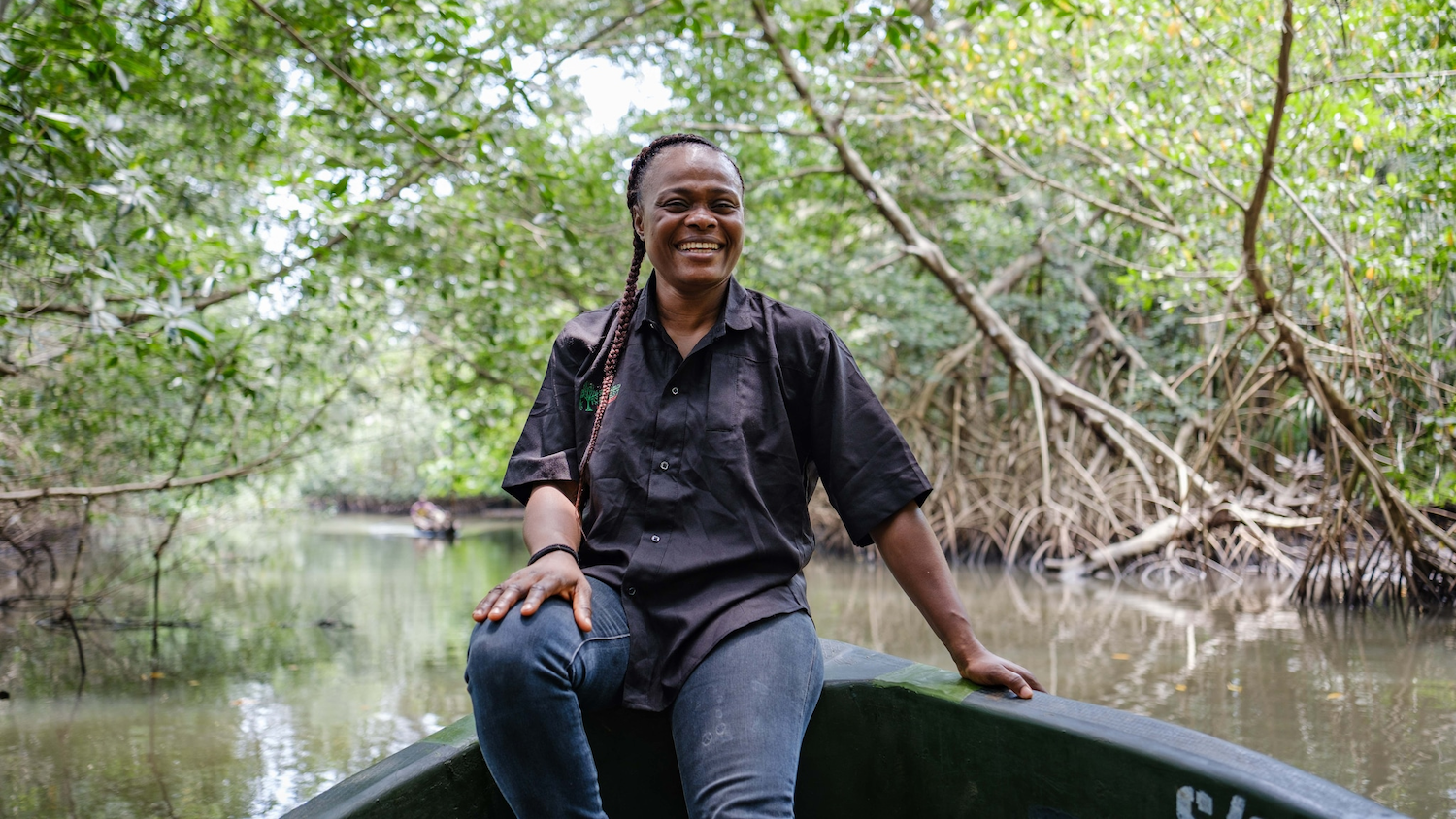 Personne souriante en bateau dans une mangrove, entourée d'arbres et d'eau calme.