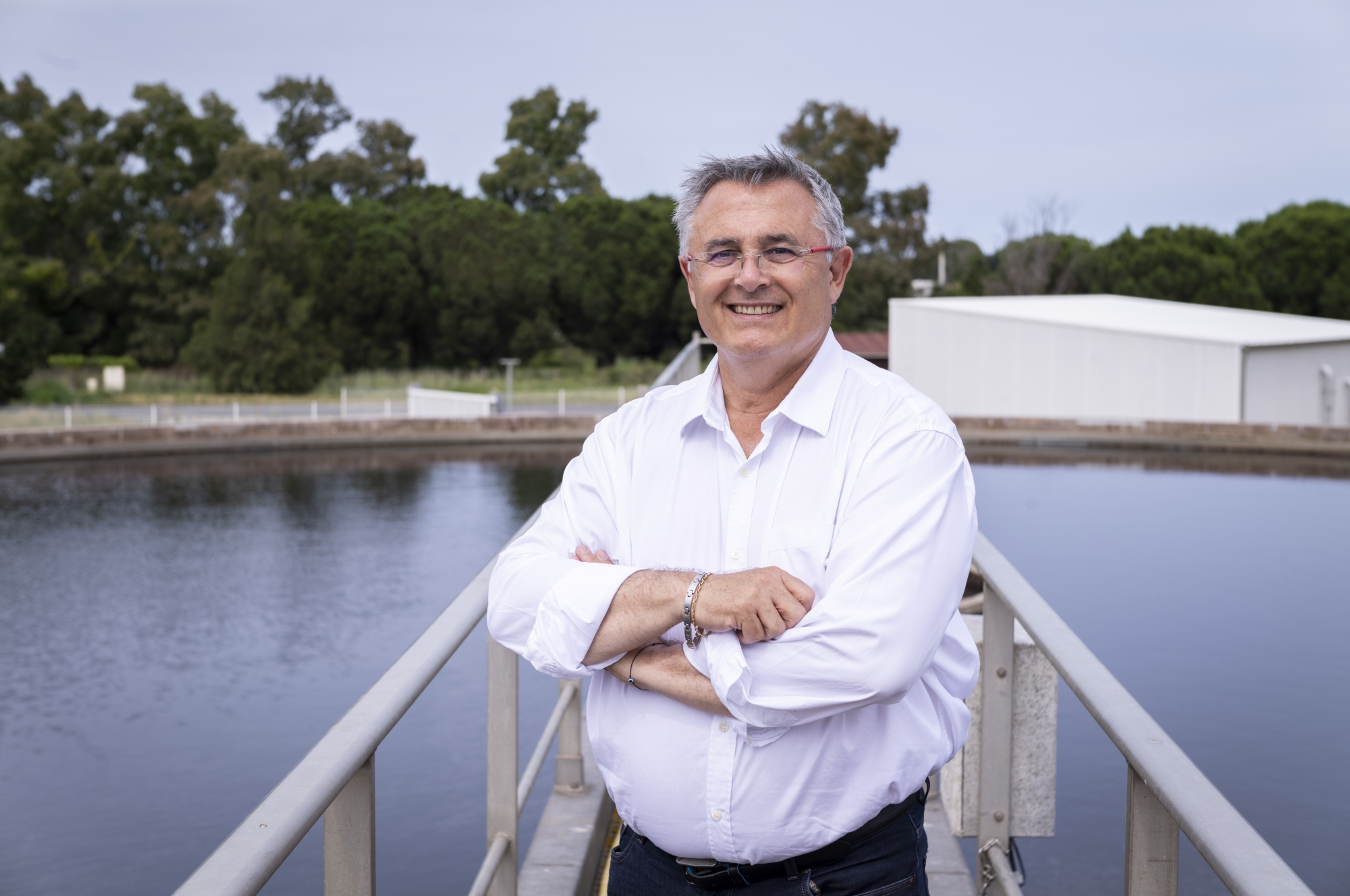 Homme souriant en chemise blanche devant un bassin d'eau et des arbres.