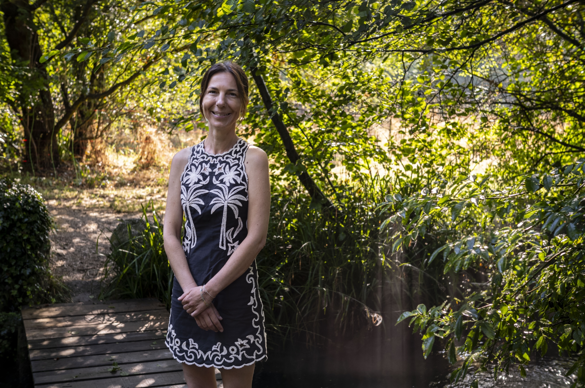 Femme souriante en robe noire avec motifs, debout dans un jardin verdoyant.
