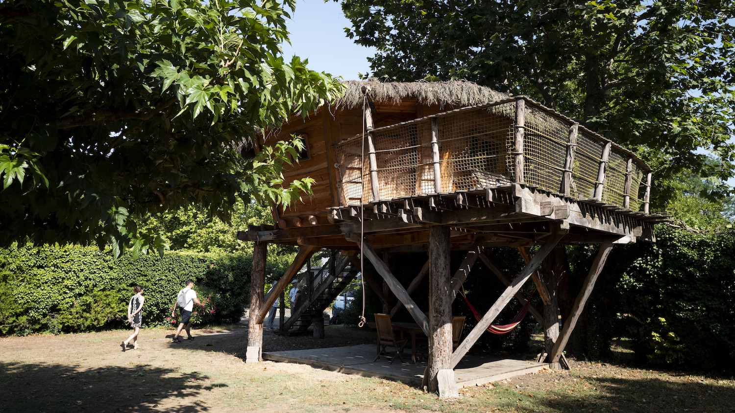 Cabane en bois sur pilotis entourée d'arbres, avec des personnes et vélos à proximité.