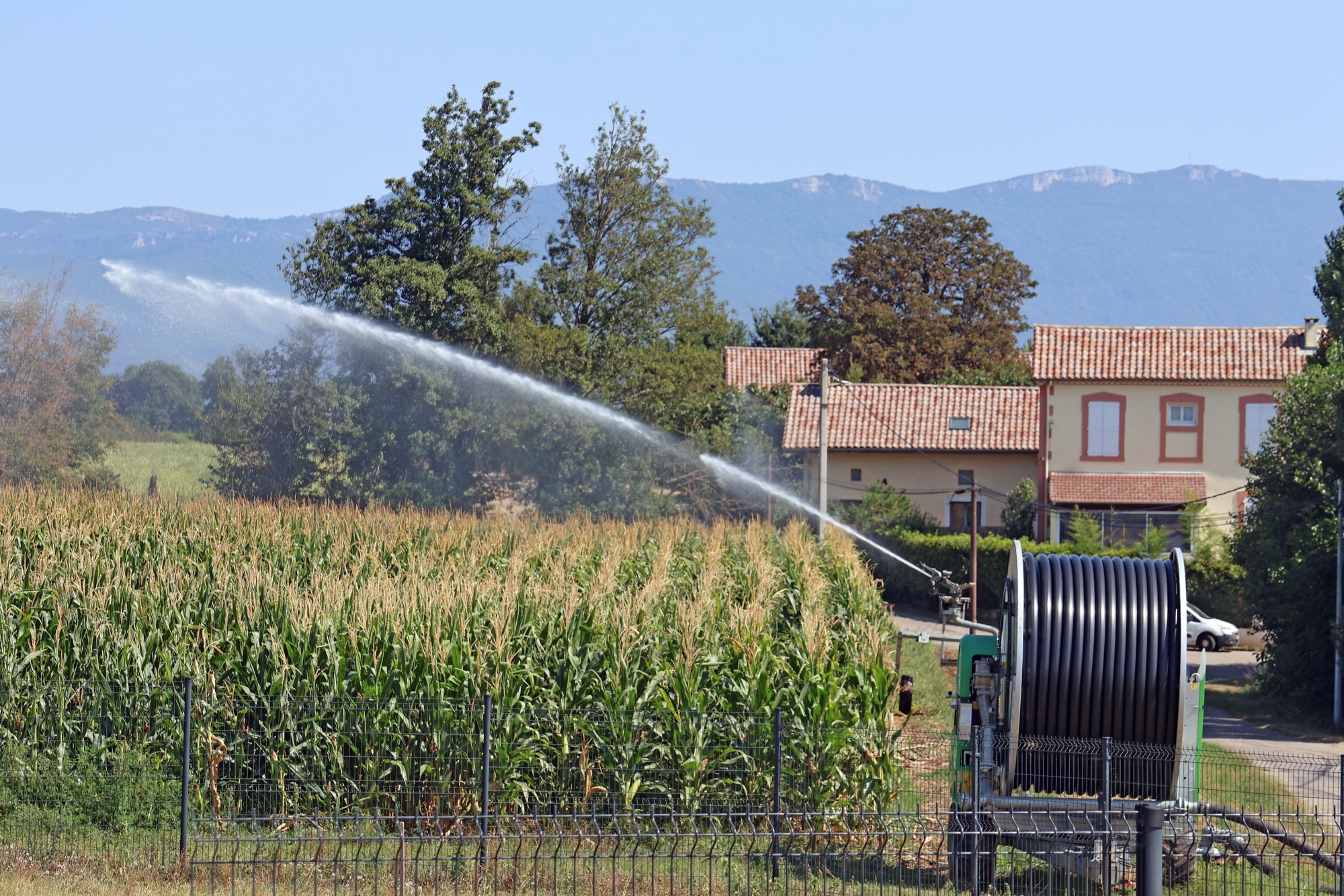 Système d'irrigation arrosant un champ de maïs près d'une maison avec montagnes en arrière-plan.