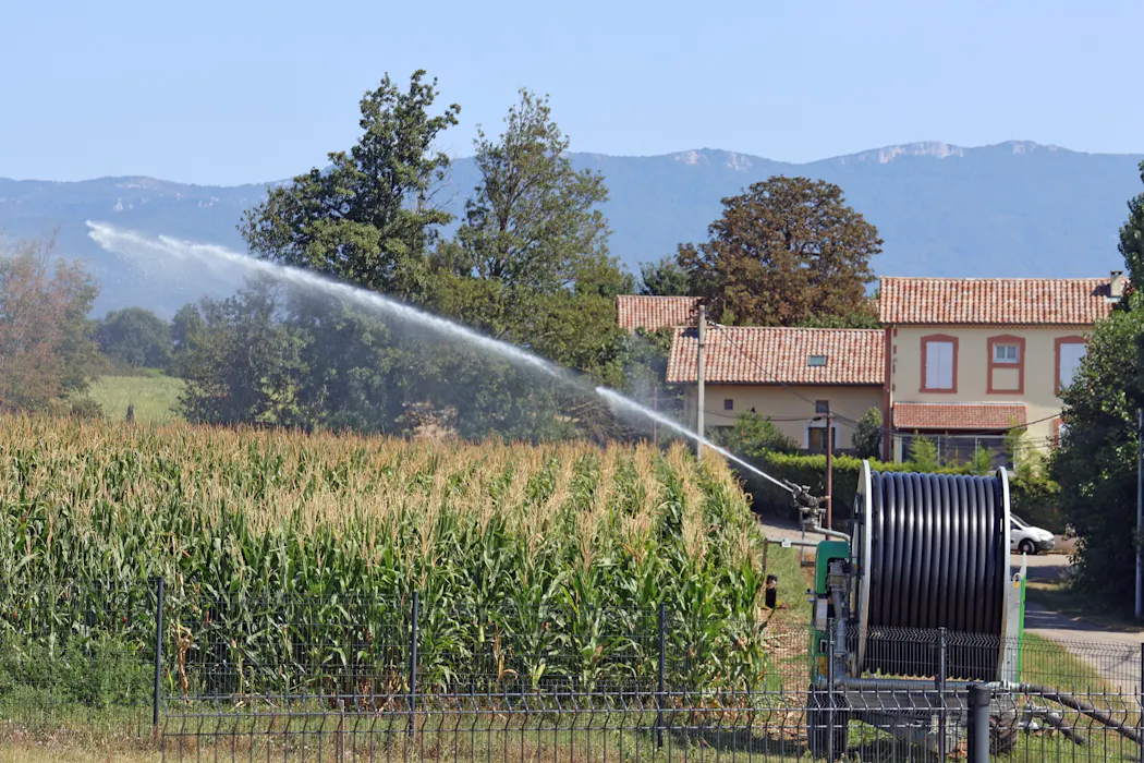 Système d'irrigation arrosant un champ de maïs près d'une maison avec montagnes en arrière-plan.