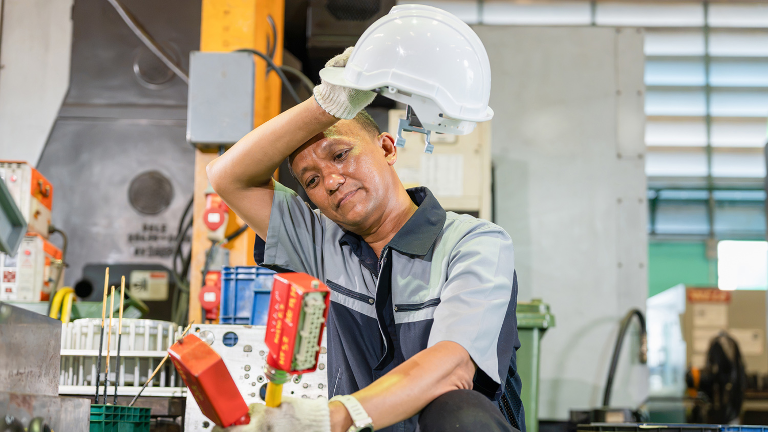 Un ouvrier fatigué s'essuie le front dans une usine, tenant un casque de sécurité.
