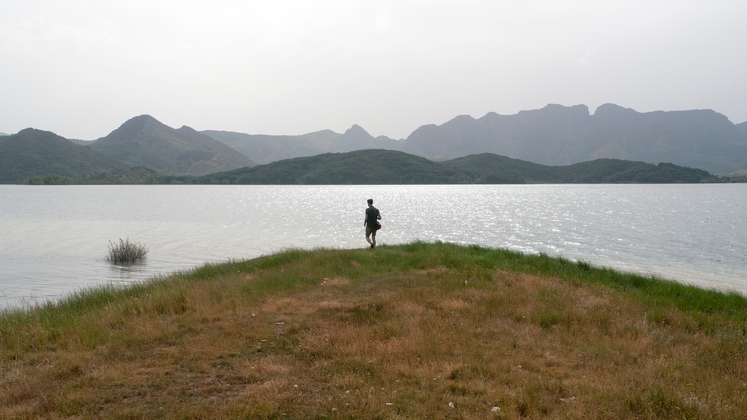 Une personne marche vers un lac entouré de montagnes sous un ciel gris.