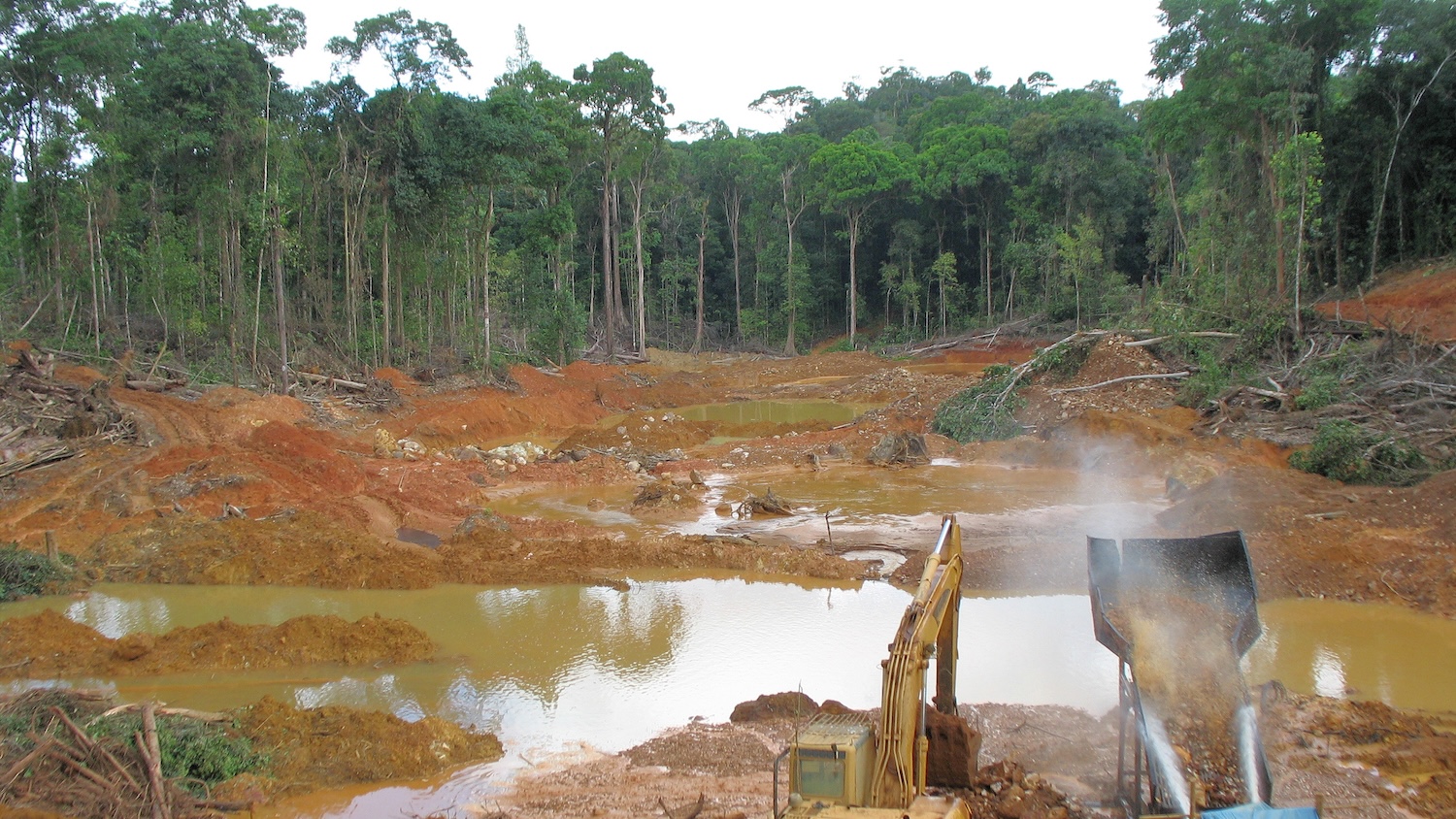Déforestation en Amazonie avec excavatrice, terre rouge et arbres abattus près d'un bassin d'eau.