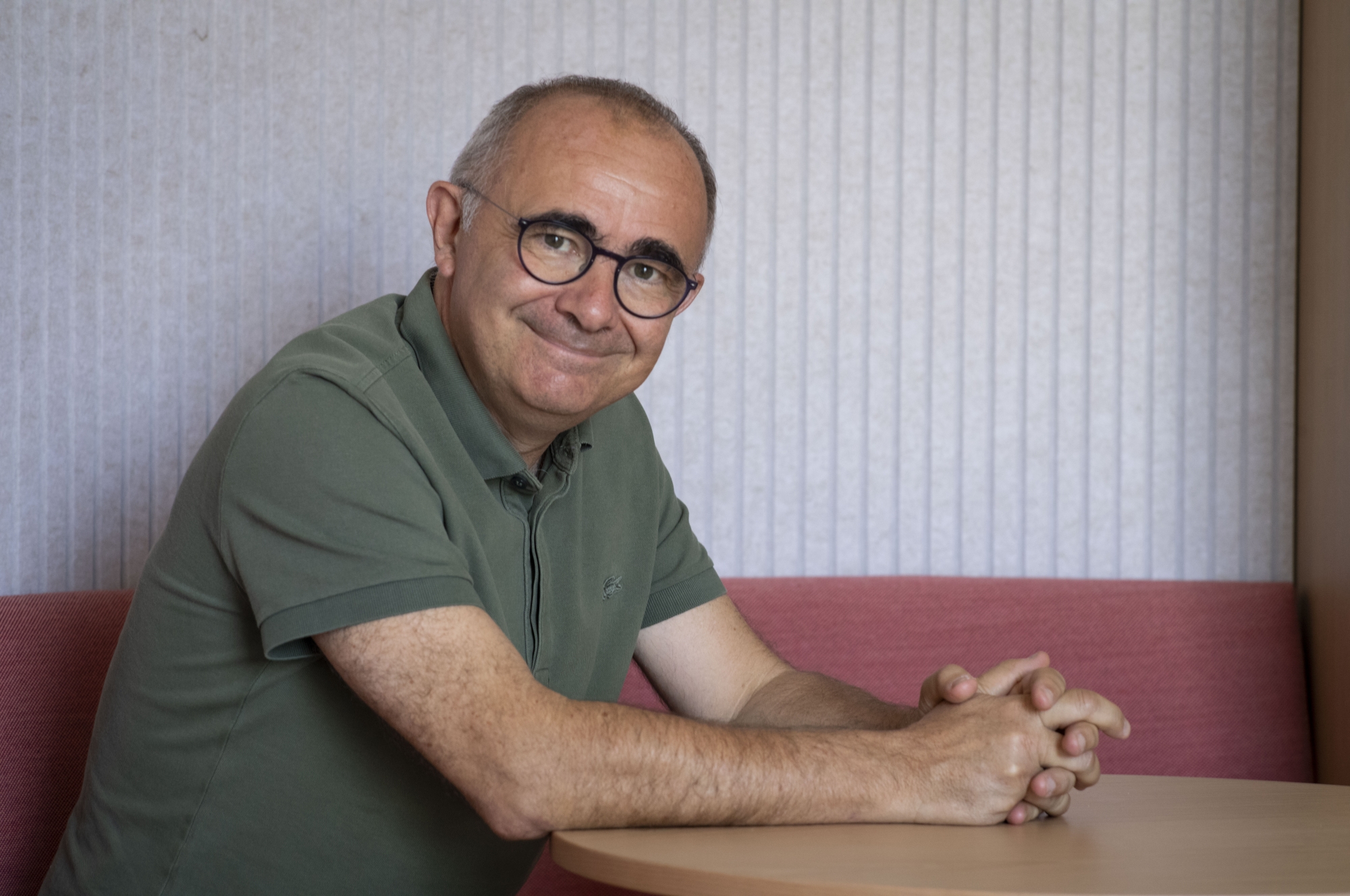 Homme souriant assis sur un canapé, portant des lunettes et un polo vert.