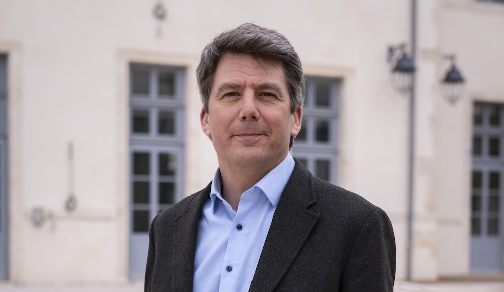 Homme en costume devant un bâtiment avec fenêtres et lampadaires, en extérieur.