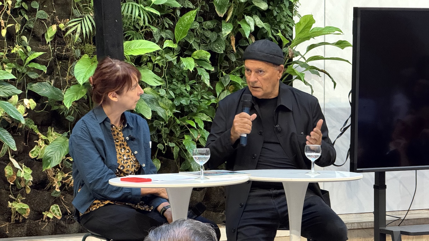 Deux personnes discutant lors d'un événement, avec plantes en arrière-plan et table ronde.