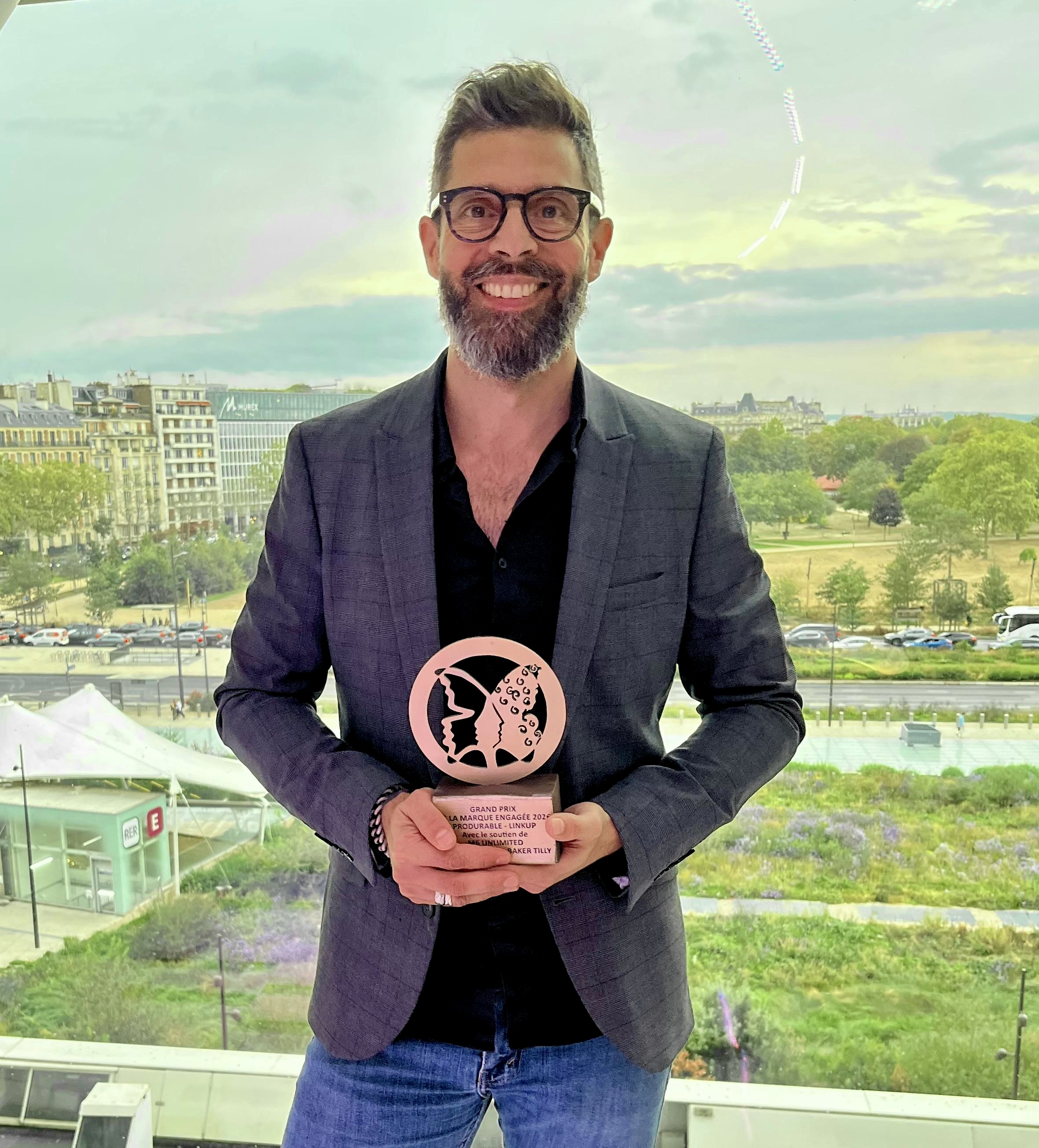 Homme souriant avec barbe et lunettes, en costume, fond de paysage urbain et ciel nuageux.