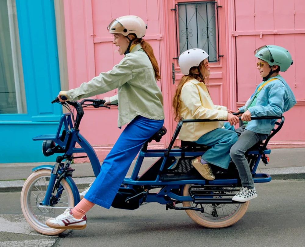 Une femme avec deux enfants sur un vélo cargo devant des murs colorés.